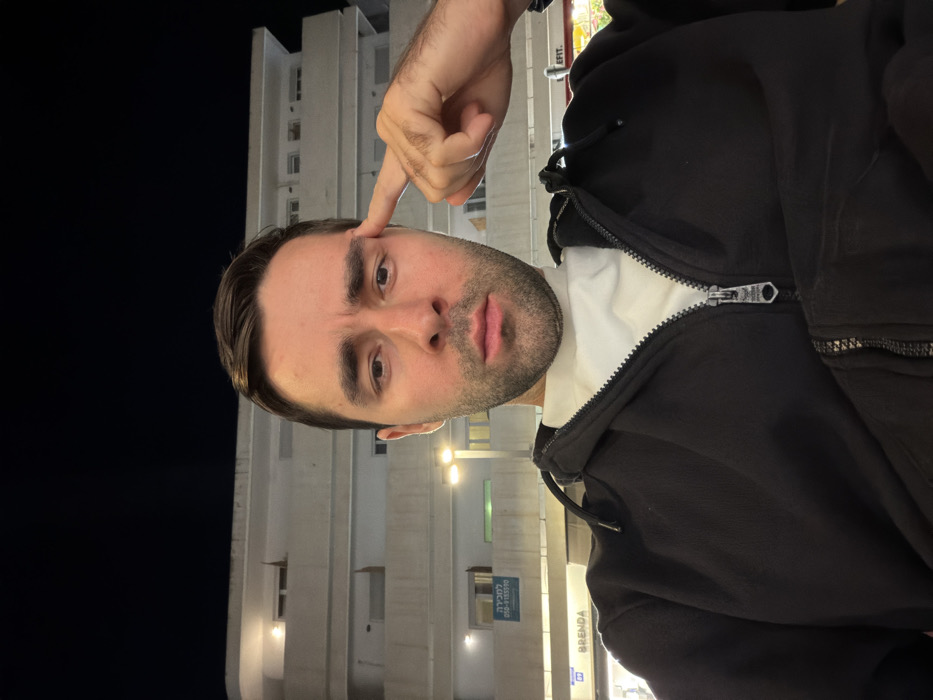 A person faces forward in a vertically oriented selfie. The person is wearing a black zip-up jacket over a white shirt. They are pointing to their temple with a raised index finger. The background includes a light-colored building with windows and a sign with visible text. The sky is dark, suggesting it is nighttime. Artificial lighting illuminates the building and the person's face.