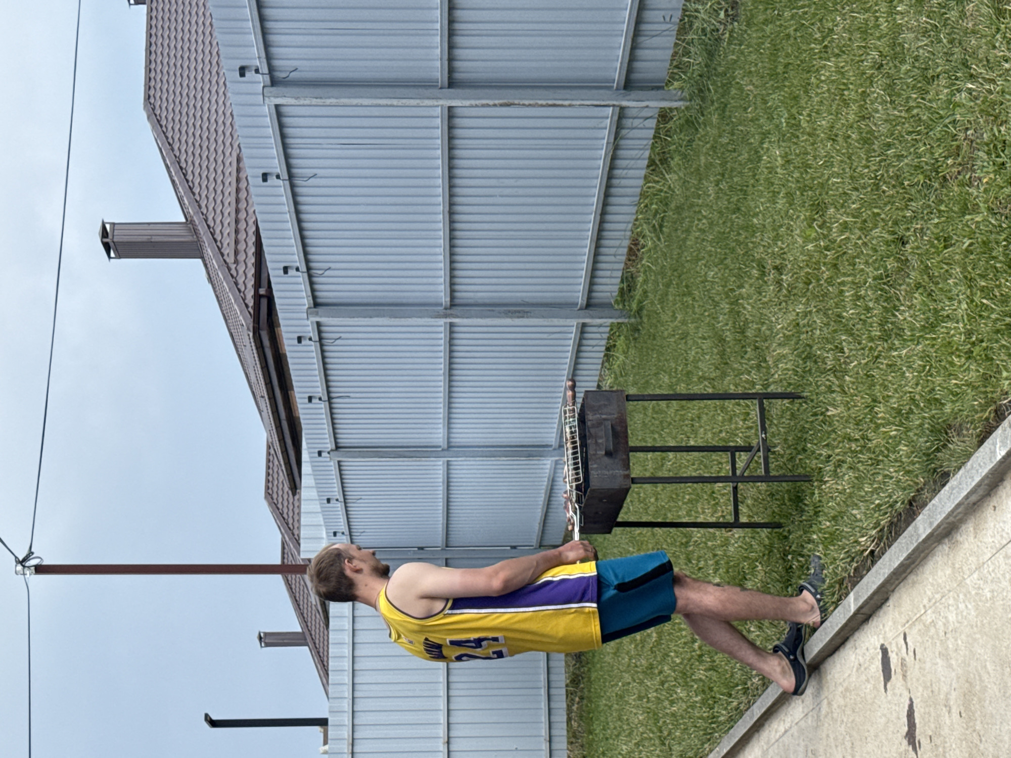In the village of Dëmino, Russia, a young man wearing a yellow and purple basketball jersey with the number 24 and blue shorts stands in a grassy yard, tending to a barbecue grill. The grill, a dark metal rectangular box on a stand, holds a metal rack with food items. A light gray corrugated metal fence runs horizontally across the background, with a portion of a brown-roofed house visible behind it. The sky is overcast. The man is wearing dark clogs on his feet. The yard is covered in green grass, with a concrete path running along the left side of the image.