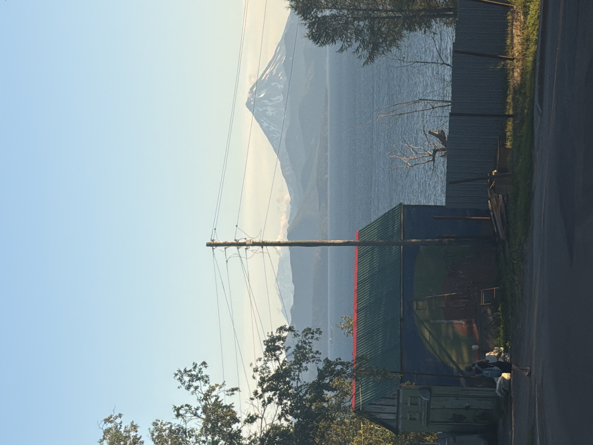 This image captures a view of a body of water with a snow-capped volcano in the background, likely Avachinsky or Koryaksky volcano, visible from Petropavlovsk-Kamchatsky, Russia. In the foreground, a small, dilapidated building with a green metal roof and a blue-painted wall featuring a mural of a dome shape is present. A person with a backpack is walking away from the building towards the viewer, wearing a dark top and shorts. A utility pole with wires runs horizontally across the scene, partially obscuring the view of the water and the distant mountains. A corrugated metal fence runs along the edge of the property. The sky is clear and blue, suggesting it is daytime, possibly morning or late afternoon given the lighting. The overall scene depicts a rural or semi-urban setting with a striking natural landscape.