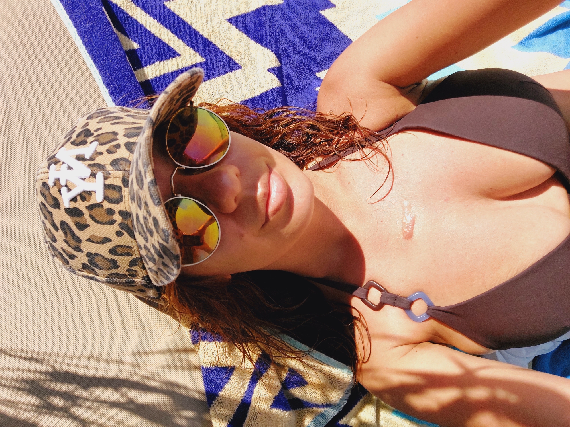 In an outdoor setting, a woman with auburn hair, wearing a brown bikini top, round sunglasses with reflective lenses, and a leopard-print baseball cap with a white "YM" logo, lies on a striped blue and white towel. The background consists of a textured beige surface and a hint of palm tree shadows. The woman's head is tilted back slightly as she looks towards the camera. The lighting suggests a sunny day. The provided information about Sozopol, Bulgaria is not visible in the image.