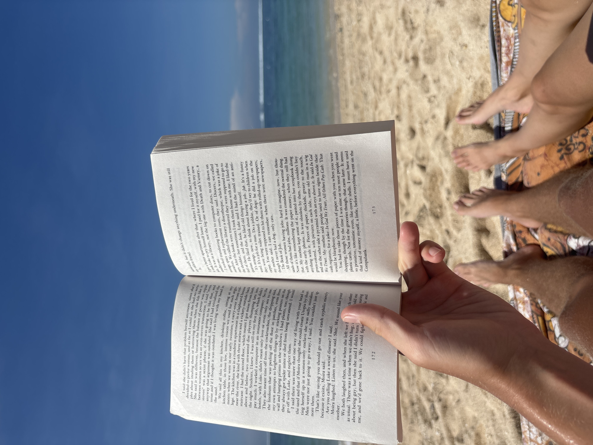 The photograph, likely taken in Kuta Selatan, Indonesia, shows a person holding an open book on a beach. The beach is sandy, with a visible shoreline. The sky is clear and blue, indicating a sunny day. In the upper right, there are at least two people sitting on a blanket or towel, with parts of their legs and feet visible. The open book is held in the foreground, obscuring some of the background. The book's pages are filled with text in English. The overall composition suggests a relaxed beach scene with someone reading a book.