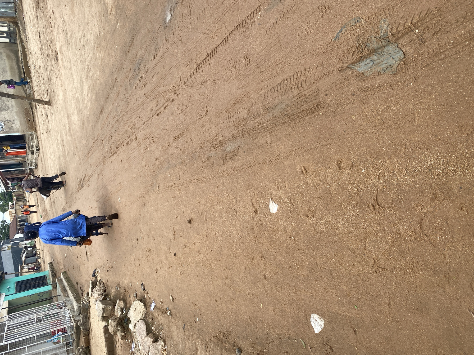 This media file shows an outdoor scene in Jalingo, Nigeria, likely a street or unpaved road. The ground is sandy and dry, with visible tire tracks from vehicles, indicating recent activity. There are several people present, some of whom are riding on motorcycles. One person in the foreground is wearing a bright blue long-sleeved shirt and is seated on a motorcycle, facing away from the camera. Another motorcycle with two riders is visible further down the path, moving away from the viewer. Several buildings, appearing to be shops or homes, line the side of the road. Some of these buildings have corrugated metal roofs and concrete walls. There are also some piles of rocks and debris on the side of the path. The overall impression is of a bustling, everyday scene in a semi-urban or rural Nigerian setting. The lighting suggests it is daytime, but the time is not precisely determinable. The weather appears dry. No text is clearly visible in the image.
