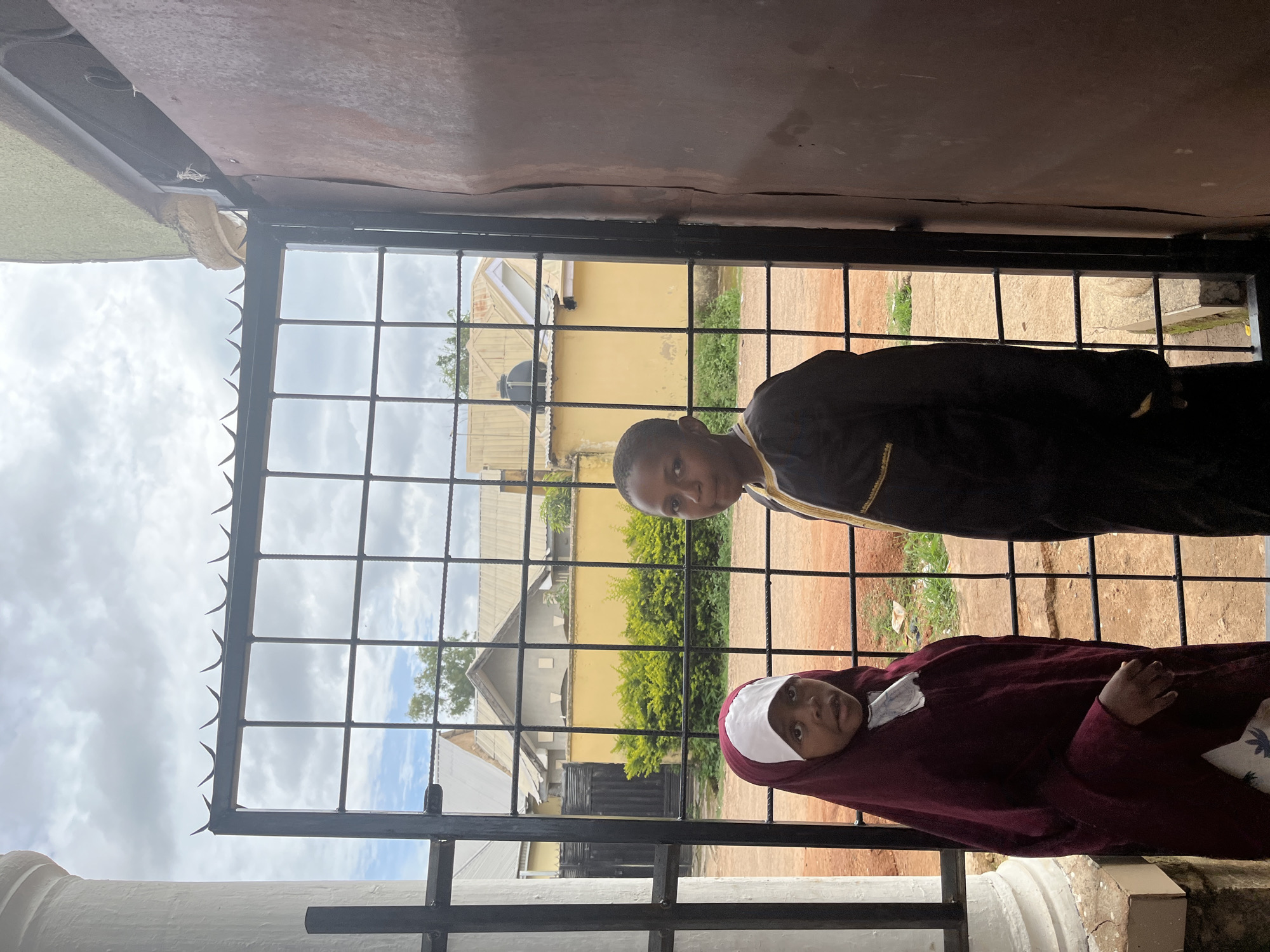 The scene, located in the city of Jos, Nigeria, depicts two children standing behind a black metal security gate. The gate is constructed with a grid pattern of reinforcing bars and is topped with a series of sharp, curved anti-climb spikes. On the left, a young girl wears a maroon hijab over a white cap, with a white garment visible at her chest; she looks directly forward. To her right, a second child, possibly a boy, wears a dark, long-sleeved garment adorned with yellow or gold trim around the collar, also gazing towards the viewer. Both children have dark skin tones. To the far left of the frame, a segment of a white, fluted architectural column is visible, suggesting a formal entrance. To the far right, a weathered, reddish-brown corrugated metal or wooden panel forms part of an adjacent wall or door. The background features a residential street with several single-story buildings under an overcast sky. These buildings display light-colored roofs, one of which supports a black water tank. A yellow-painted wall with green shrubbery in front of it is visible directly behind the children. The ground visible through the gate consists of reddish-brown earth with scattered patches of green vegetation.