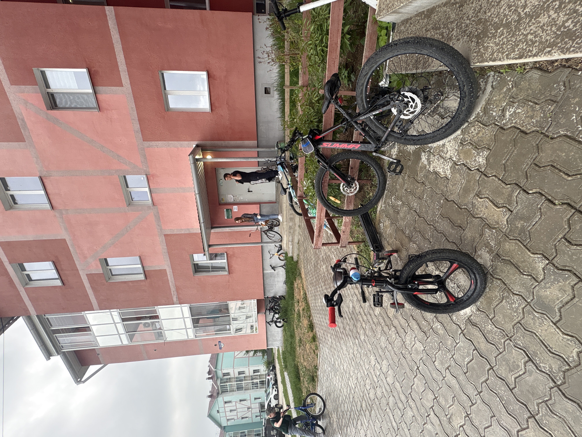 The image depicts a building exterior on a gray, possibly overcast day. The building is a light red/salmon color and appears to be multi-story, with several windows visible. In front of the building, on a paved area of interlocking blocks, are two bicycles. The bike in the foreground is black and red, while the other is black. Near the building entrance, a few people are visible, including one person standing in the doorway. The scene appears to be in an urban area, as other buildings are visible in the background. The location is identified as Birobidzhan, Russia.