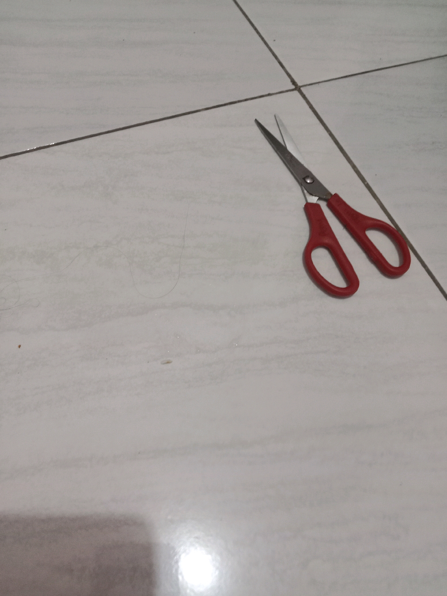 A pair of red-handled scissors is positioned on a tiled floor. The scissors are open, with their blades pointing upwards and slightly to the left. The floor is a neutral color with visible grout lines. There is a shadow in the lower portion of the image. The image is taken from a high-angle perspective. The location is not specified in the image or its context.