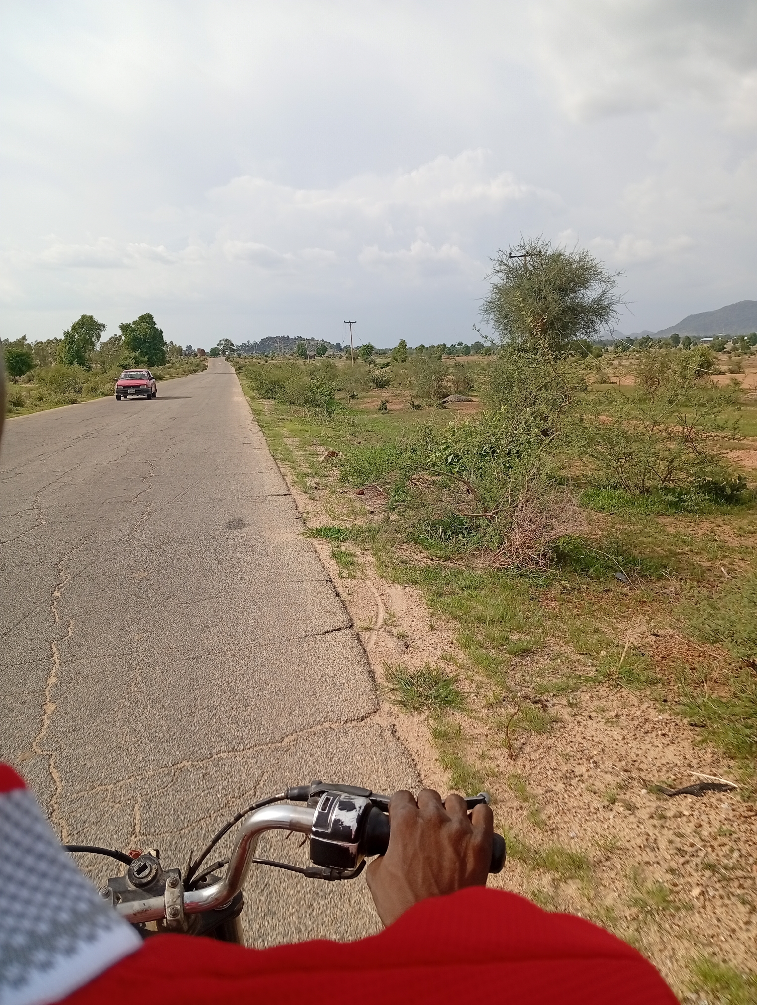 The image shows a view from the perspective of someone riding a motorcycle on a road in Shuwa, Nigeria. The road is asphalt and appears to be in moderate condition, with visible cracks. To the left of the road, a red car is visible in the distance, driving in the same direction. The landscape on either side of the road is rural, characterized by sparse vegetation, scattered trees, and dry, sandy soil. In the background, hills are visible, and the sky is overcast with clouds. A power pole with wires is also present on the right side of the road. The handlebars and a portion of the rider's arm and red clothing are in the foreground.