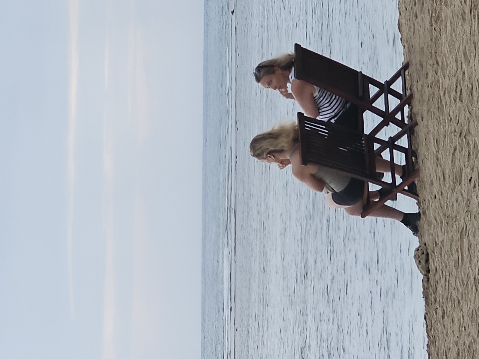 Two women are sitting in lounge chairs on a sandy beach, facing the ocean. The woman on the left has blonde hair and is wearing a dark tank top and shorts. The woman on the right has blonde hair and is wearing a striped tank top. The ocean is calm with gentle waves, and the sky is overcast. The scene is likely a beach in Denpasar, Indonesia, given the tropical setting and typical beach activities. The women appear relaxed and are looking out at the water, possibly enjoying the view or having a conversation. The lighting suggests it is either early morning or late afternoon.