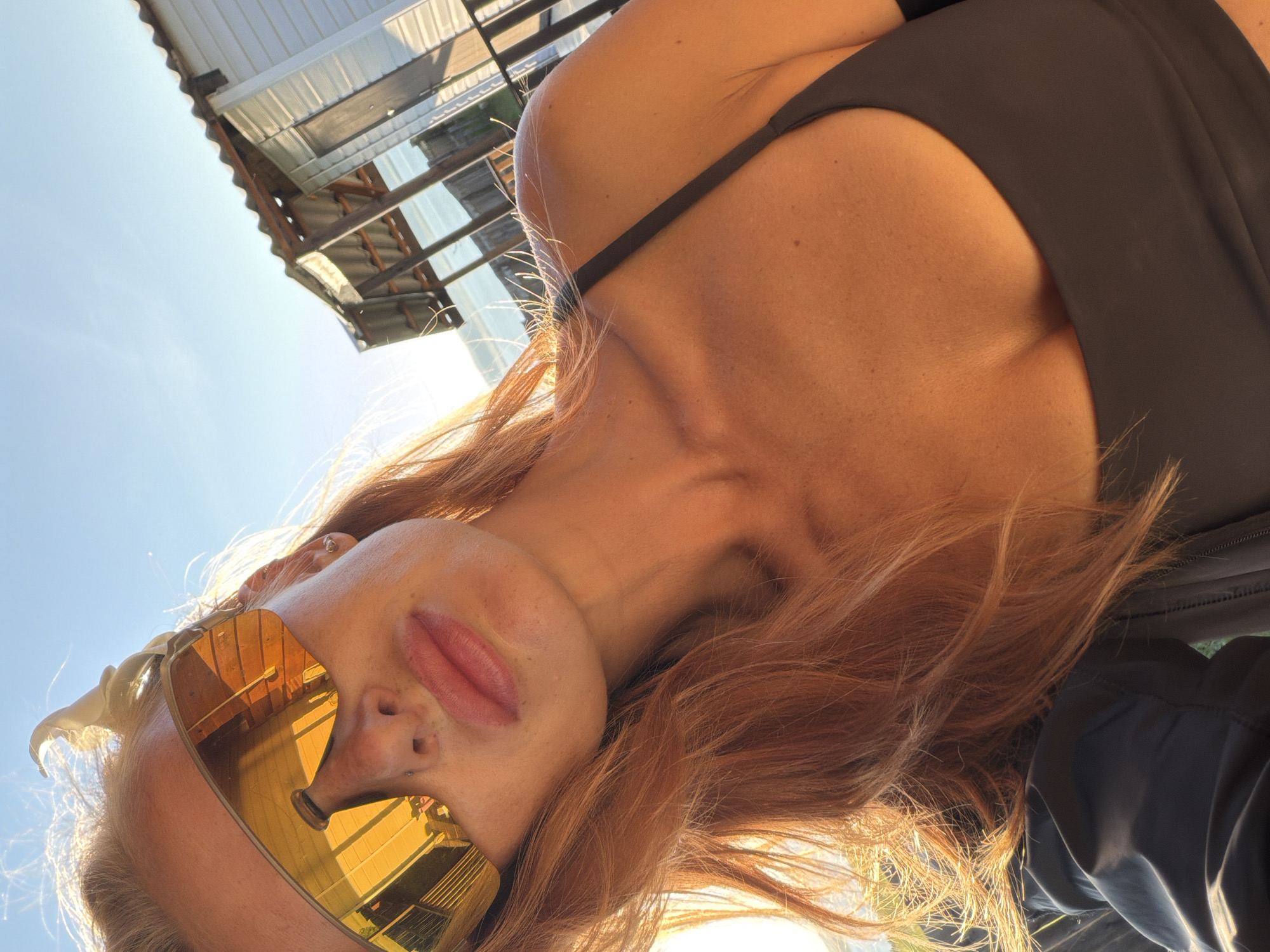 In an outdoor selfie taken in an unknown location in Russia, a young woman with reddish-blonde hair and prominent freckles poses against a bright blue sky. She is wearing large, mirrored sunglasses with an orange tint, a dark-colored bikini top, and a dark jacket. Her hair is windswept, with strands catching the sunlight. In the background, a small, white wooden structure with a corrugated metal roof is visible, suggesting a rustic or coastal setting. The image is characterized by strong sunlight, creating a warm, golden hue across the woman's skin and hair.