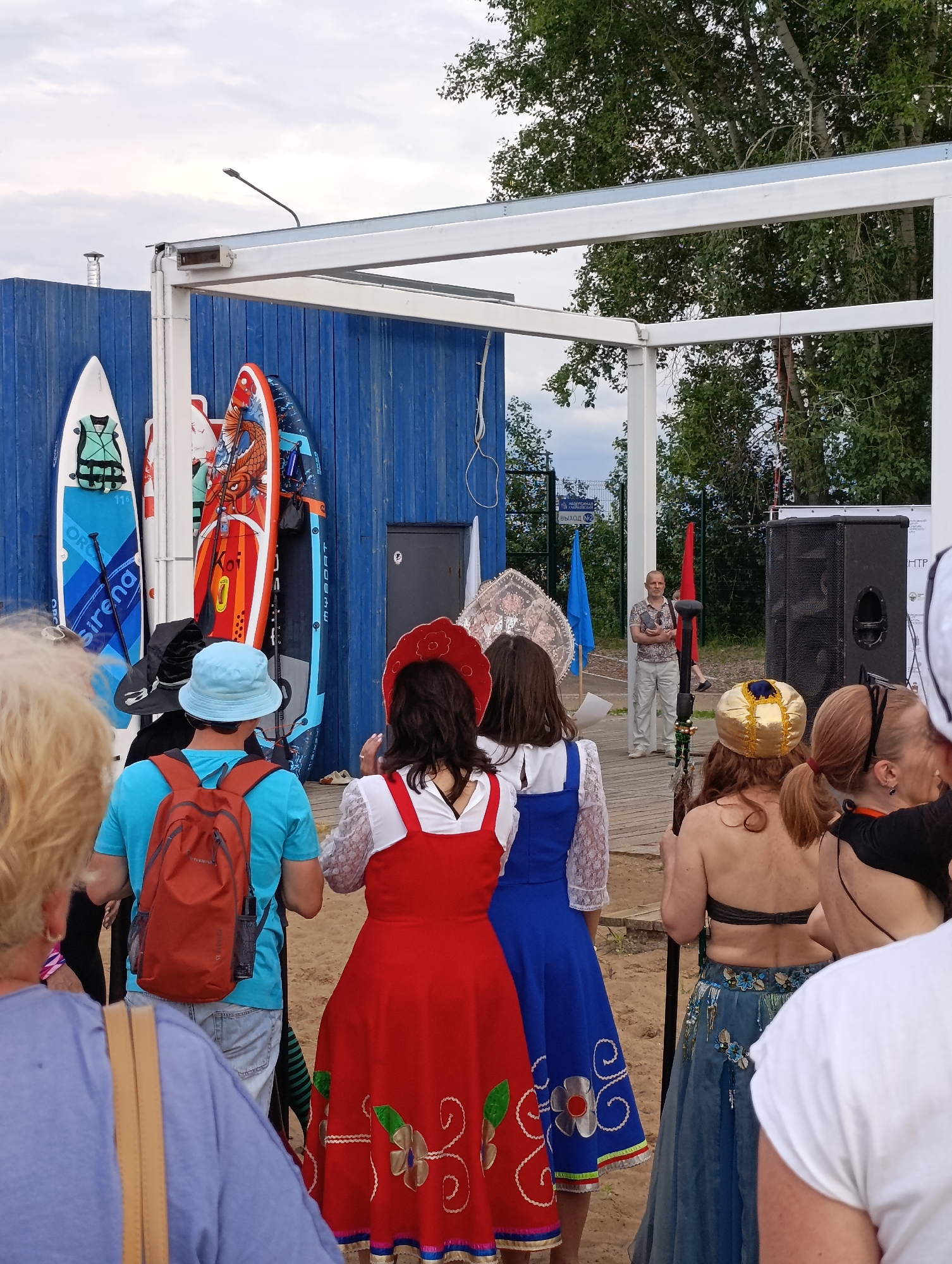 The image depicts an outdoor scene at what appears to be a beach or waterfront area in the city of Chaikovsky, Russia. In the background, a vibrant blue wooden structure is visible, adorned with several paddleboards leaning against it. These boards feature colorful graphics, including one with a koi fish design. A white, open-air canopy with metal framing extends over a portion of the scene.
In the foreground and mid-ground, a diverse group of people are gathered. Several individuals are facing away from the camera, some wearing traditional-style clothing. One woman is dressed in a red folk-inspired dress with intricate floral embroidery. Another person is wearing a blue dress with similar decorative elements. A man in a blue t-shirt and a light blue hat with an orange backpack is also present.
Further back, a man in a patterned shirt and light-colored pants stands near a large black speaker. Two flags, one blue and one red, are visible behind him. The ground appears to be sandy. Trees with green foliage are visible in the upper portion of the image, suggesting a natural environment. The sky is overcast. The overall impression is of a community gathering or event taking place in an outdoor recreational setting.