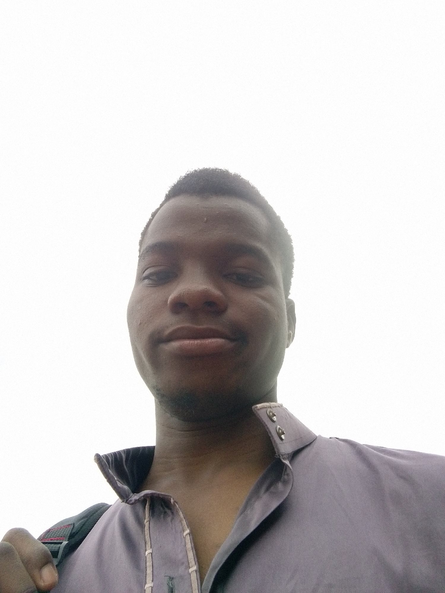 A low-angle close-up selfie of a dark-skinned man with short, dark hair and a slight beard. He is wearing a lavender shirt with a high collar and two visible silver buttons. Part of a backpack strap is visible over his left shoulder. The background is a bright, overcast sky. No specific details indicate the location is Eleweran, Nigeria; this is not confirmed.