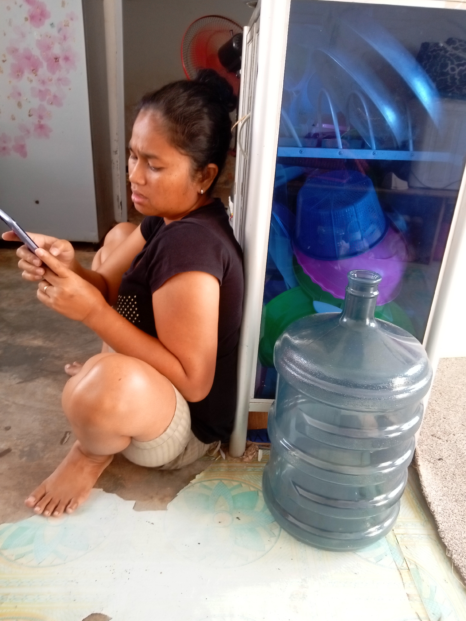 The image depicts a woman sitting on the floor, looking at her mobile phone. She is wearing a black t-shirt and light-colored shorts. Her legs are crossed, and her right hand is holding the phone while her left hand is resting on her lap. Beside her is a large, empty water jug. Behind the woman is a white cabinet with a blue glass door, revealing stacked dishes and colorful plastic containers inside. A red electric fan is visible behind the cabinet. The floor appears to be tiled with a patterned design, and a small section of a rug is visible in the lower right corner. The overall environment suggests an indoor or semi-outdoor setting, possibly a home or a small shop, in Batam, Indonesia.