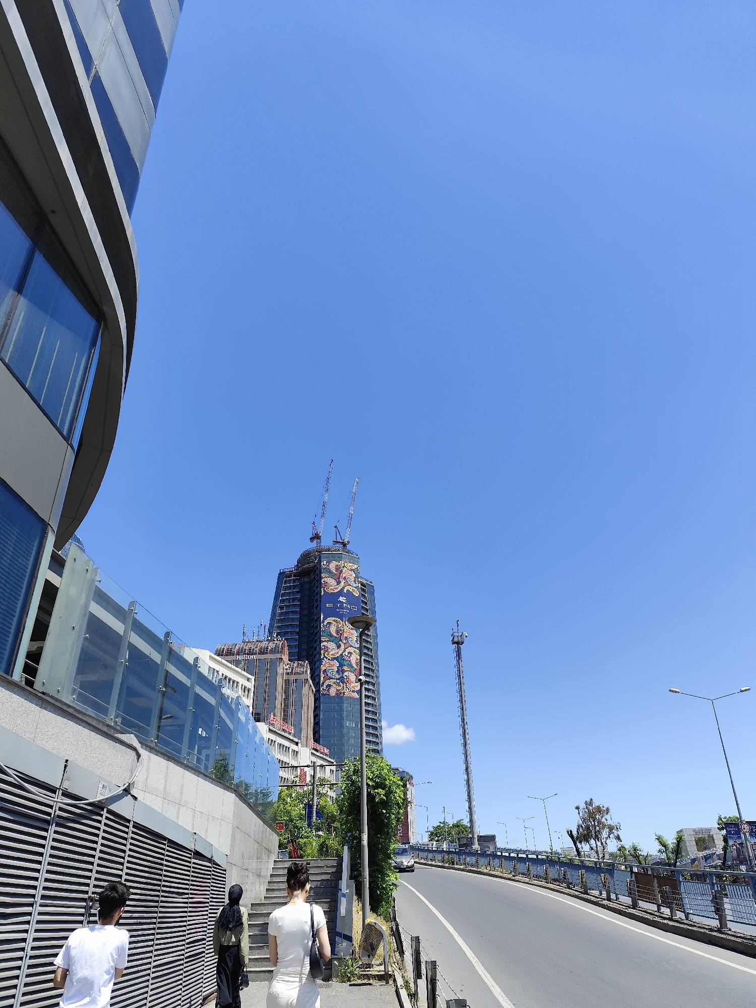 Here is a forensic description of the image:
The image shows an urban street scene in Sarıyer, Turkey, under a clear blue sky. On the left, part of a modern building with curved glass windows is visible. In the foreground, three people are walking away from the viewer on a sidewalk. The first person is wearing a white t-shirt, the second is wearing a black hijab and a light green top, and the third person is wearing a white dress. Next to the sidewalk, there are steps leading to an entrance, partially obscured by metal bars.
In the background, a tall skyscraper under construction is wrapped in a large advertisement. Several other buildings, including one with "Hilton" signage, are visible. Two cranes top the skyscraper. To the right, a communication tower stands tall. A road stretches into the distance, bordered by a blue railing. Streetlights line the road, and some vegetation is present along the roadside. A car is seen driving on the road.