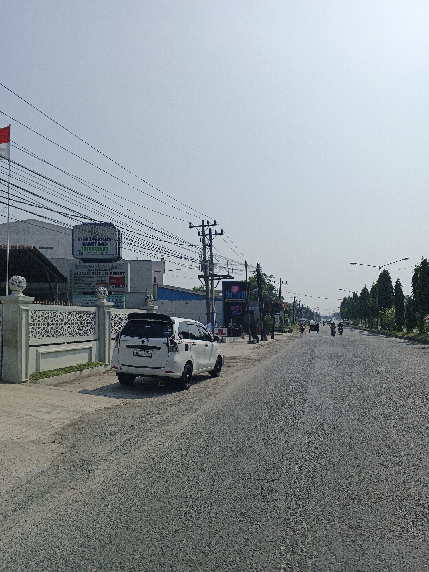 The image depicts a street scene in Tg Morawa B, Indonesia, on a clear day with a bright sky. The foreground is dominated by a paved road with visible gravel and texture. On the left side of the road, a white car is parked on the side. Adjacent to the car is a building with a white decorative fence and a sign that reads "KLINIK PERTAMA RAWAT INAP TUTUN SEHATI" and "FASKES TINGKAT 1 KLINIK TUTUN SEHATI". There are several utility poles with a dense network of wires running across the sky. In the middle ground, the road stretches into the distance, with several motorcycles and a few cars driving on it. On the right side of the road, a row of tall, slender trees are planted along the sidewalk. Further down the road, more buildings and signs are visible, including a sign for "GOGASIN". The overall environment appears to be a mix of commercial and residential areas.