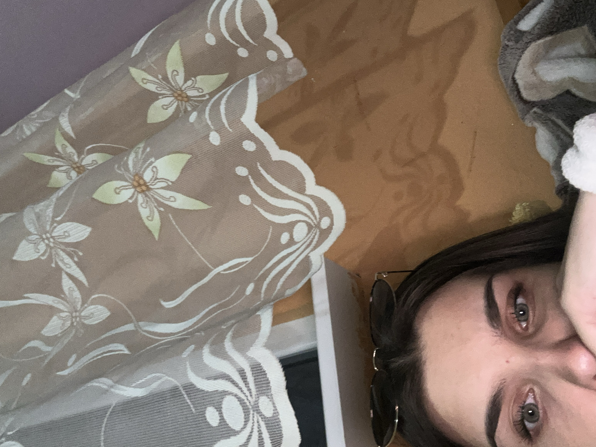 The image is a close-up, indoor, selfie-style photograph. The subject is a young person with dark hair, wearing sunglasses perched on their head. Their eyes are open and looking towards the camera, and their right hand is partially covering their mouth. The background features a sheer curtain with a floral pattern, appearing to be behind a window or on a wall. Below the curtain, a wooden surface is visible, possibly a wall or furniture. A patterned blanket or throw is also visible in the lower right corner of the frame. The lighting suggests an indoor setting, possibly during the day, with some ambient light. There are no clear indicators of the city of Shumen, Bulgaria, or any specific outdoor environment.