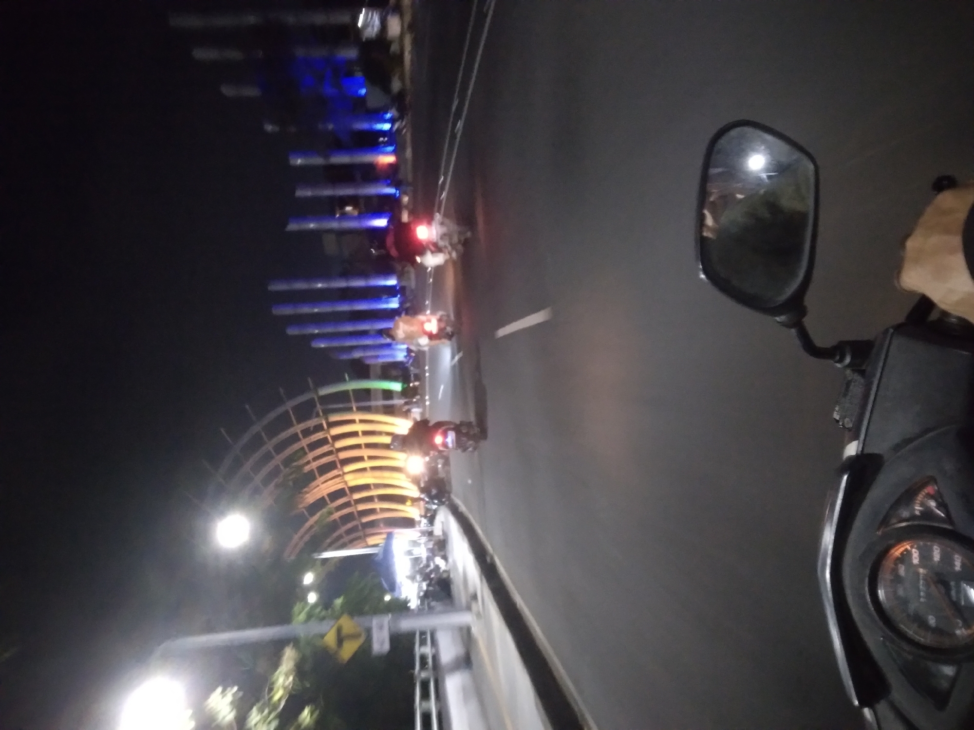 The image captures a nighttime view from the perspective of someone riding a motorcycle on a paved road in Purwokerto, Indonesia. The road has white lane markings and is bordered by a concrete barrier and sidewalk on the left. On the left side of the road, there is a street lamp and a yellow diamond-shaped road sign with a black border and a black 'T' symbol. Further down the sidewalk, a blue tent is visible with motorcycles parked around it.
The background features a striking illuminated architectural structure with a curved, ribbed design, lit by warm orange and yellow lights. Beyond this, a series of tall, slender vertical structures are illuminated with vibrant blue lights, suggesting a modern urban or public space.
Several motorcycles are on the road. In the middle ground, three motorcycles are traveling in the same direction as the viewer. The closest motorcycle has its taillight illuminated red. The rider of this motorcycle is wearing a dark helmet and jacket. Further ahead, two other motorcycles are also visible, one with its taillight on. In the foreground, the motorcycle's dashboard is visible, including a speedometer with an orange needle. A rider's hand is also visible on the right side of the handlebar, gripping the throttle. A side-view mirror on the motorcycle reflects a blurry image of the rider and the surrounding lights. The overall atmosphere is one of a busy city street at night, with illuminated landmarks and vehicular traffic.