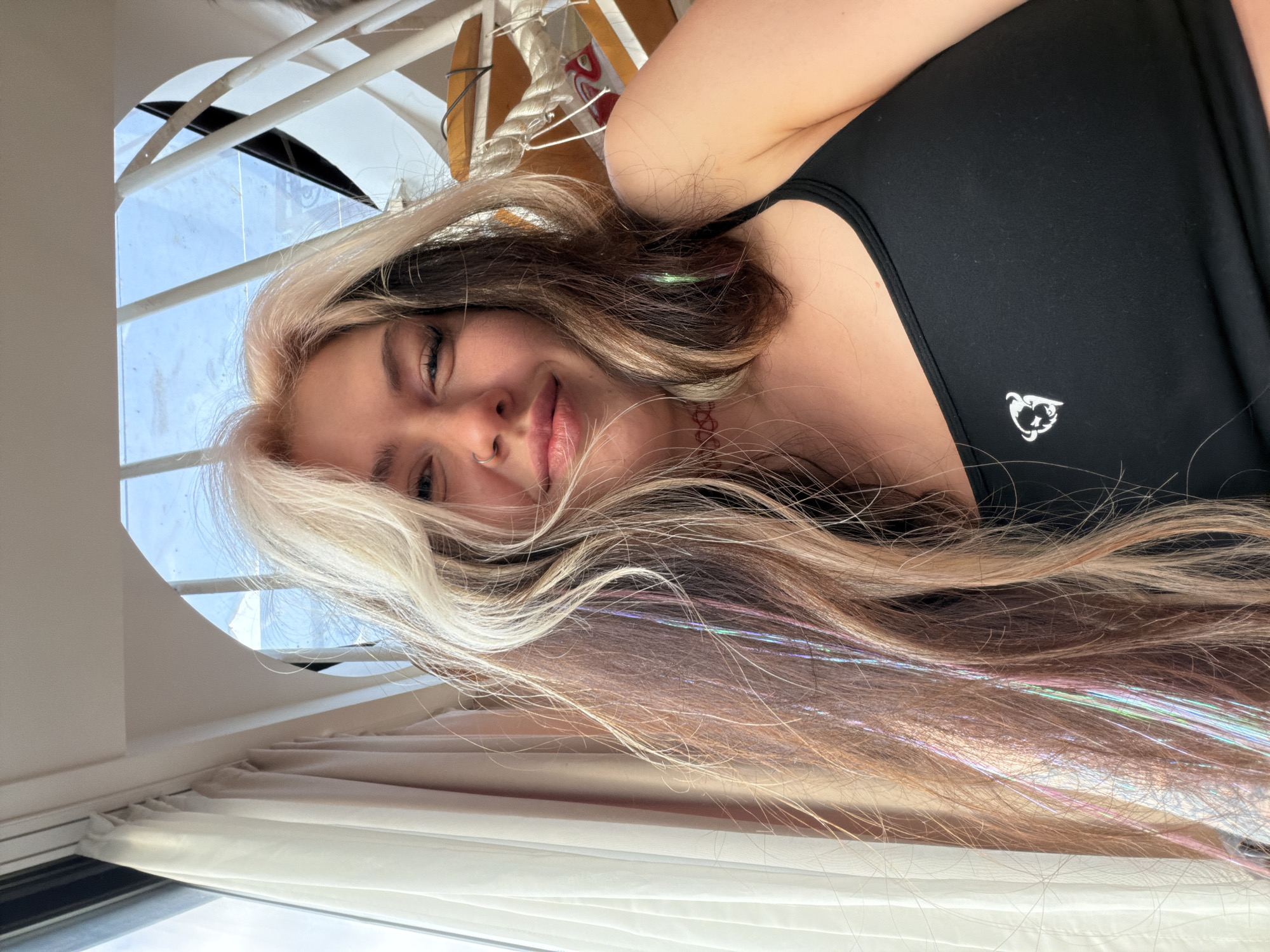 A young woman with blonde and brown hair, including hair extensions, is lying in a room with a white wall and window. She is wearing a black tank top. The location is possibly in Budva, Montenegro, based on the presence of a window and a likely indoor setting. There are white curtains and other objects visible through the window.