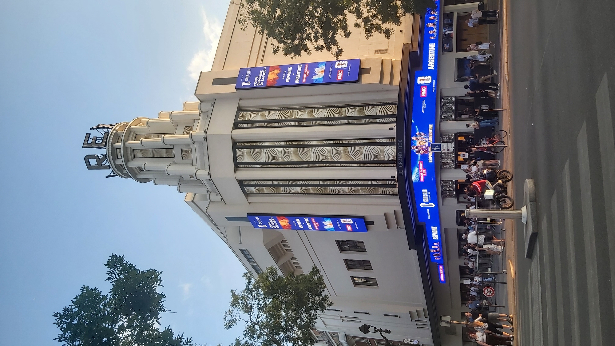 The image shows Le Grand Rex, a cinema and concert hall located in Paris, France. The building has a distinctive art deco design with a large, illuminated marquee displaying "LE GRAND REX" in blue letters. Above the marquee, banners advertise a football match final between Spain and Argentina for the FIFA World Cup. The street in front of the cinema is bustling with people, some queuing to enter and others walking by. Several vehicles, including a motorcycle and a bicycle, are visible on the street. The sky is clear and blue, suggesting it is daytime.