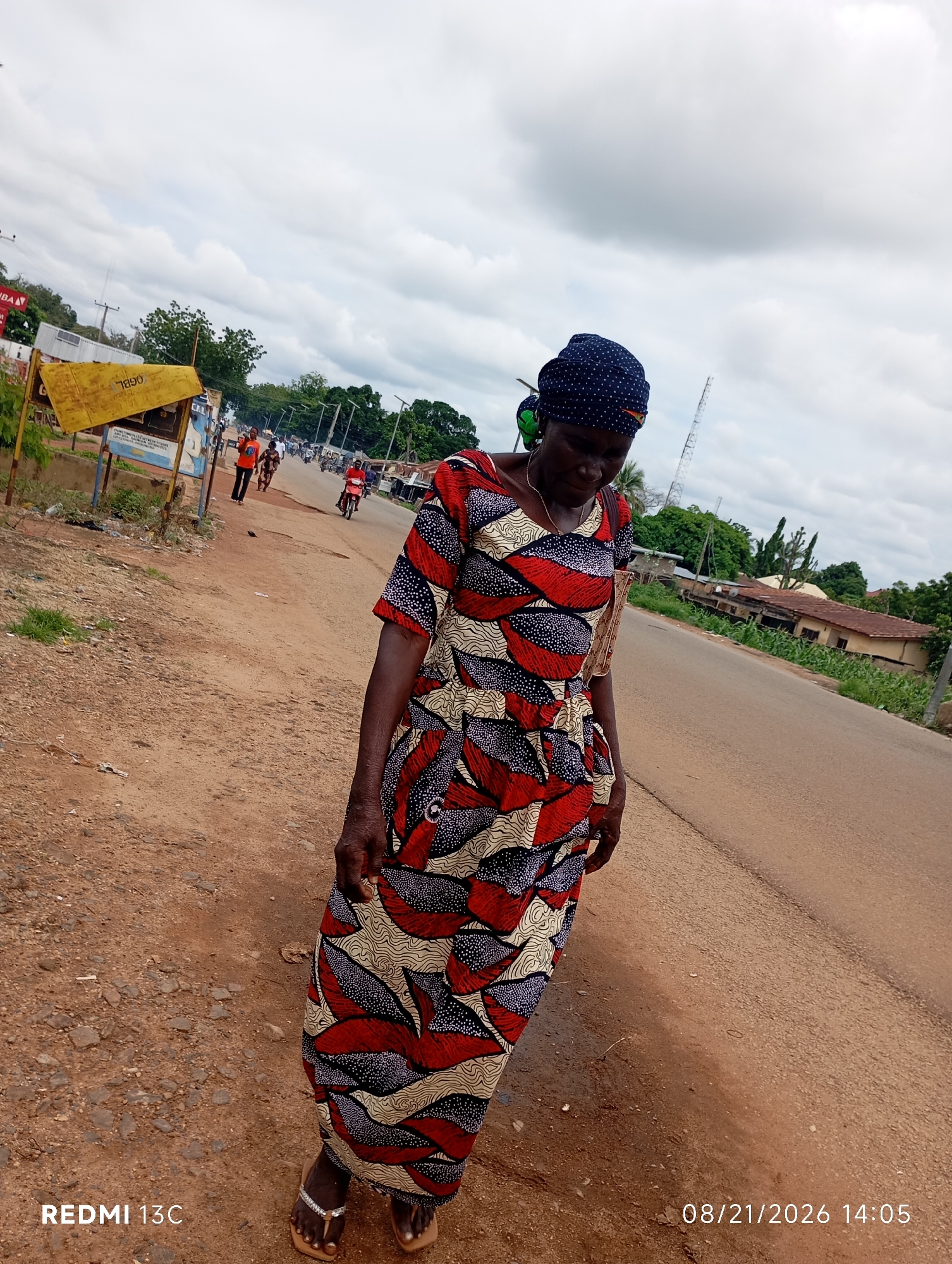 A woman wearing a blue headscarf and a colorful patterned dress walks on a dirt path next to a paved road in Shendam, Nigeria. The woman is facing downwards and to her left. In the background, there are several people walking on the road, and a motorcycle is seen in motion. Buildings and trees line the opposite side of the road. The sky is cloudy. A yellow signpost is visible on the left side of the image. The timestamp "08/21/2026 14:05" and "REDMI 13C" are superimposed on the image.