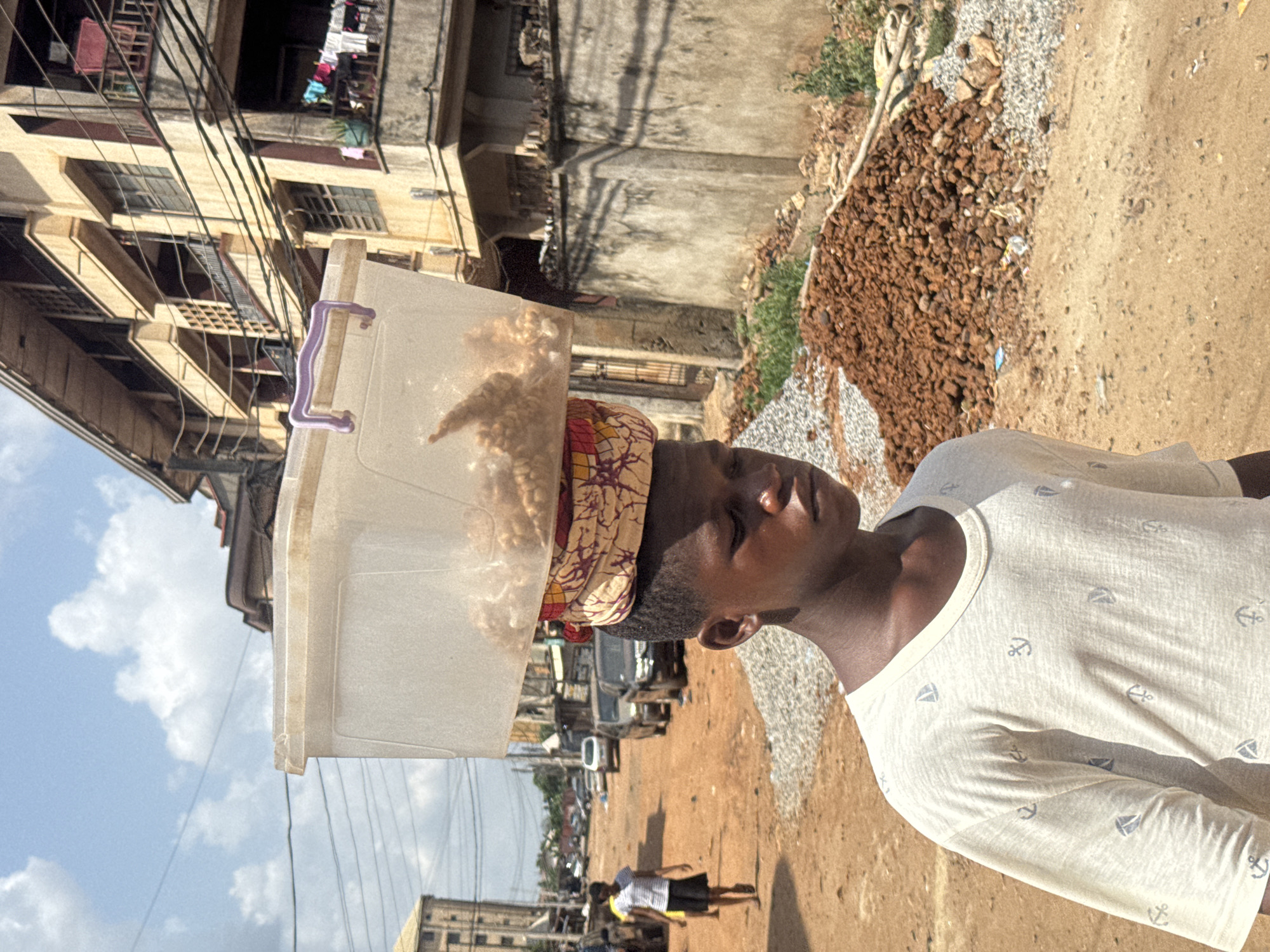 This image depicts a sunlit scene on an unpaved street in Onitsha, Nigeria. In the foreground, a young person, likely female, is central to the frame, expertly balancing a large, clear plastic storage container on their head. The container appears to hold numerous pieces of light-colored, puffed or fried snacks, suggesting they are a vendor. A vibrant headwrap, featuring yellow, red, and purple patterns, is worn as a cushion beneath the container. The individual wears a light-colored t-shirt decorated with small anchor and sailboat motifs. Their face is seen in profile, with eyes either closed or cast downwards, possibly due to the bright sun or focused on maintaining balance.
The background reveals a bustling, older urban environment. Multi-story buildings, characteristic of the region, line the left side of the frame, with dense electrical wires stretching between them. Clothes are visible hanging from some of the windows or balconies. The road surface is predominantly dirt or unpaved, with significant piles of gravel and small rocks lining the right side, indicating construction or road maintenance. Further down the street, another person is seen walking, and indistinct vehicles are present. The sky is bright blue with scattered white clouds, and the strong sunlight casts clear shadows, suggesting it is midday or early afternoon. The scene captures a common practice of head-carrying for transport and commerce in many parts of Africa. No discernible text is visible in the image.