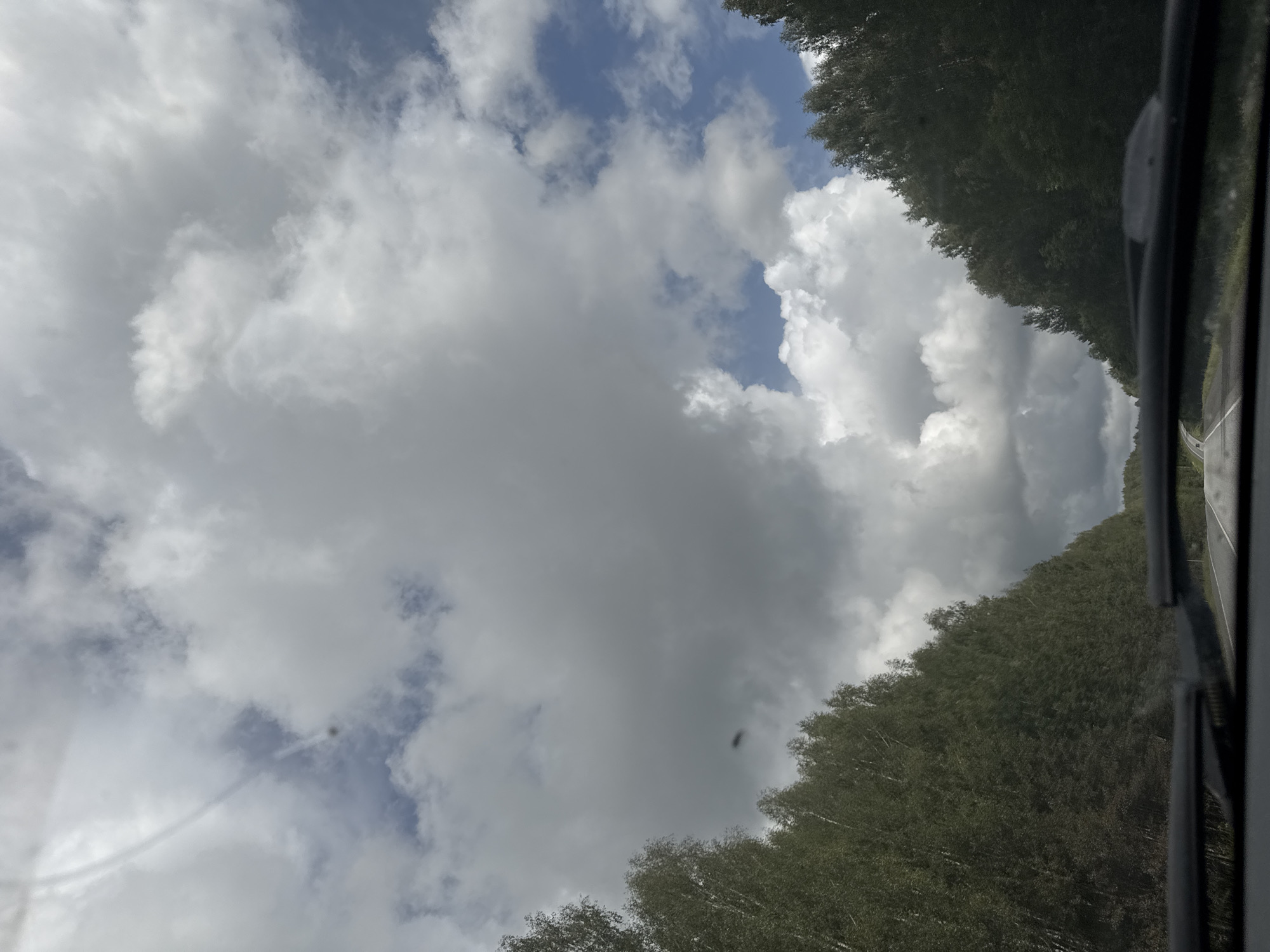 The image depicts a view from inside a vehicle, looking forward along a paved road. The road is flanked by dense trees on either side, which appear to be birch trees, creating a tunnel-like effect. The sky above is filled with dramatic clouds, ranging from dark gray to bright white, with patches of blue sky visible. The overall impression is of a drive through a forested area under a dynamic, overcast sky. The location is identified as Kazanskii Kliuch, Russia.