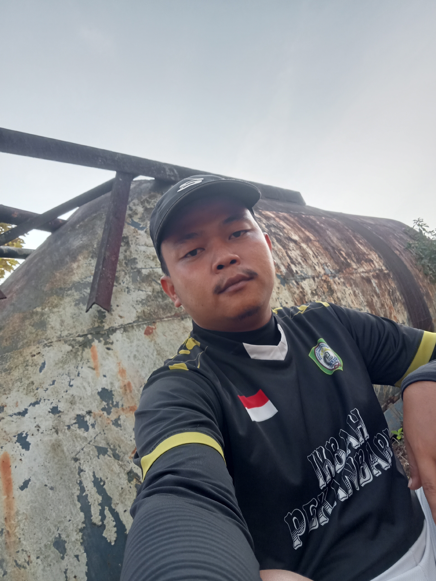 A person is taking a selfie outdoors, with a large, weathered, rusty tank in the background. The tank is a pale, chipped color with patches of rust. It appears to be a cylindrical water tank with some supporting metal structures. The sky is overcast and pale. The person is wearing a black baseball cap, a black long-sleeved shirt with yellow accents on the sleeves and collar. The shirt has a crest on the left chest, an Indonesian flag emblem on the right chest, and white lettering that reads "KBAH PEKANBAR" on the front. The person has a short mustache. The environment appears to be an outdoor, possibly industrial or derelict area, which is consistent with the location of Teluk Kenidai, Indonesia, known for its rural and industrial landscapes.
