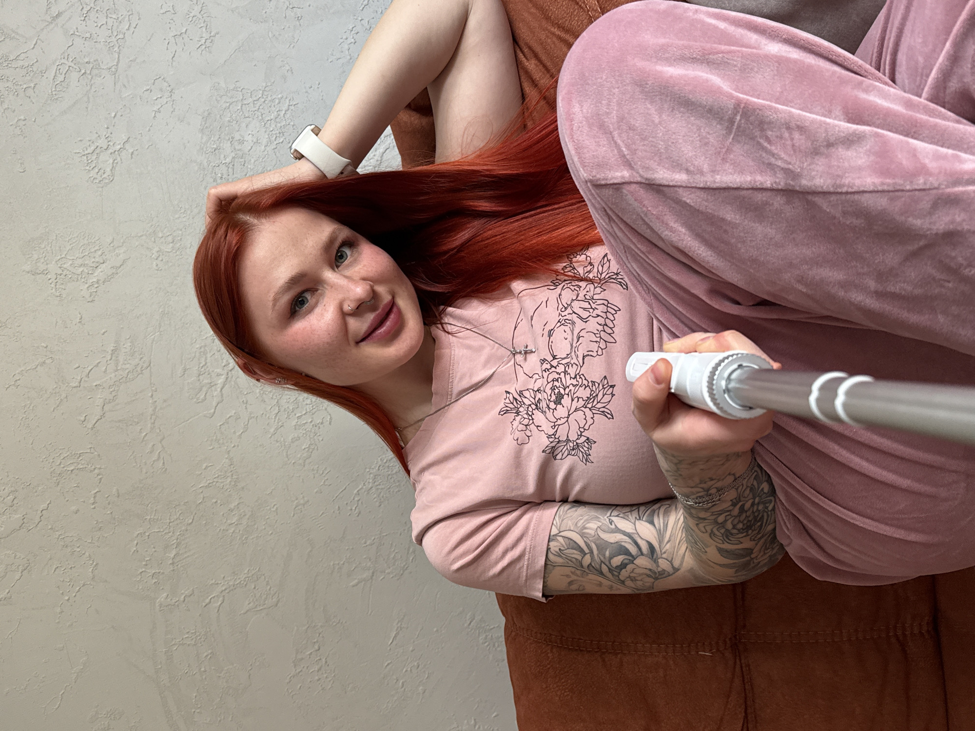 A young woman with red hair is taking a selfie while lying on an orange couch. She is wearing a pink top with a floral design and pink velour pants. She has a white smartwatch on her left wrist and a tattoo on her right arm. The background includes a light-colored textured wall. The location is Rostov-on-Don, Russia.