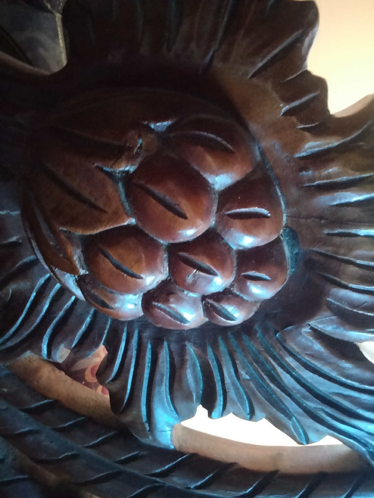 The image displays a close-up, dimly lit, and somewhat blurry view of carved dark brown wood. The carving features a cluster of bulbous shapes, each with an incised slit resembling an eye or a mouth, surrounded by petal-like or feather-like structures. The wood has a glossy finish, reflecting light in streaks of blue and white. The background is out of focus, showing warm, muted tones that suggest an indoor setting. The specific location in the city of Payagaboh, Indonesia, is not discernable from the visual information presented in the image.