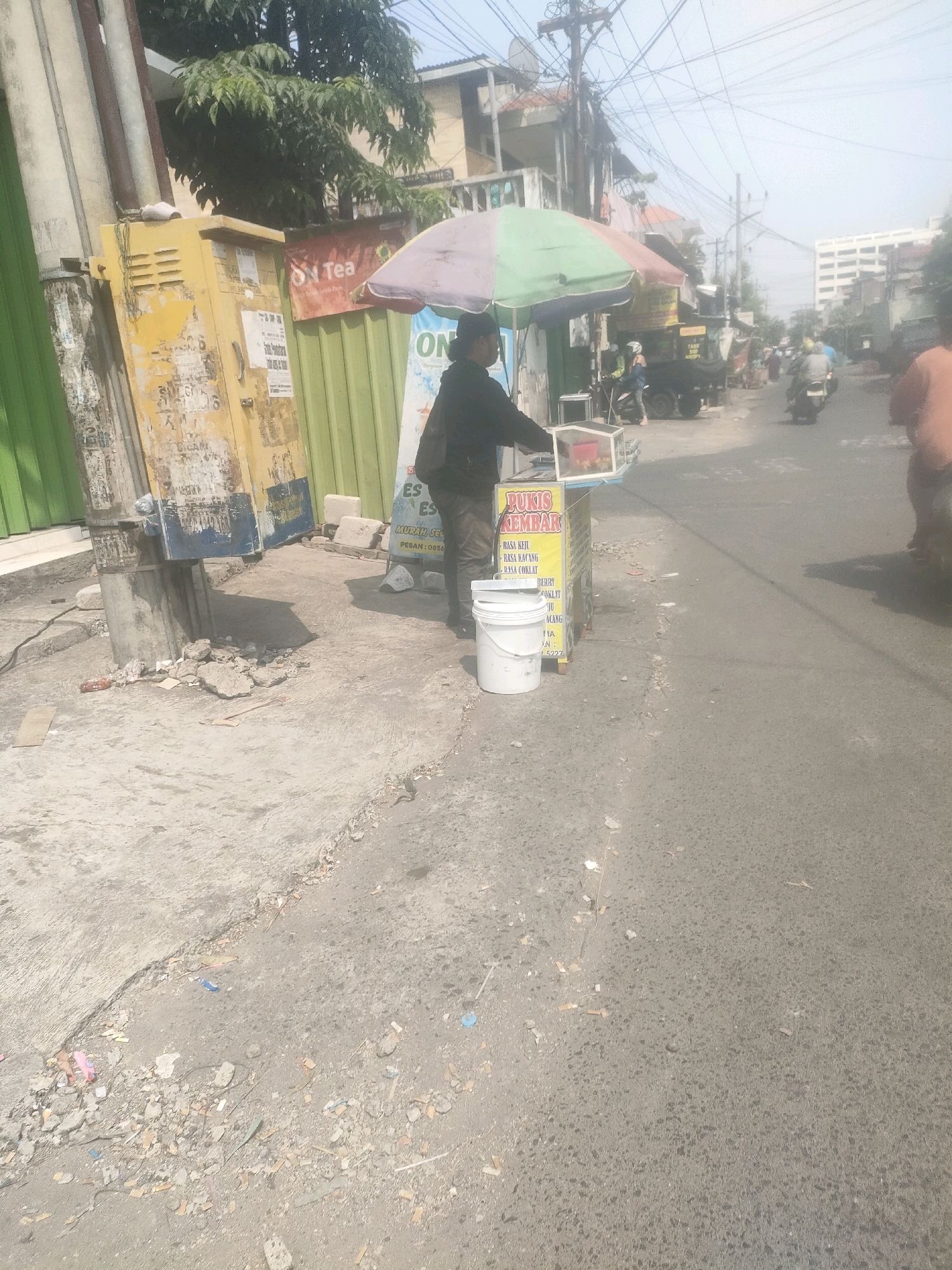 In Surabaya, Indonesia, a street vendor stands by their cart under a colorful umbrella, selling "Pukis Kembar" (twin pukis, a type of Indonesian pancake). The cart is yellow with red text, listing flavors like "Rasa Keju" (cheese), "Rasa Kacang" (peanut), and "Rasa Coklat" (chocolate). A white bucket sits beside the cart. In the background, buildings and a large white high-rise are visible. The street is paved, with some debris scattered on the ground. Several vehicles, including motorcycles and a truck, are present, indicating the busy nature of the urban environment. The sky is overcast.