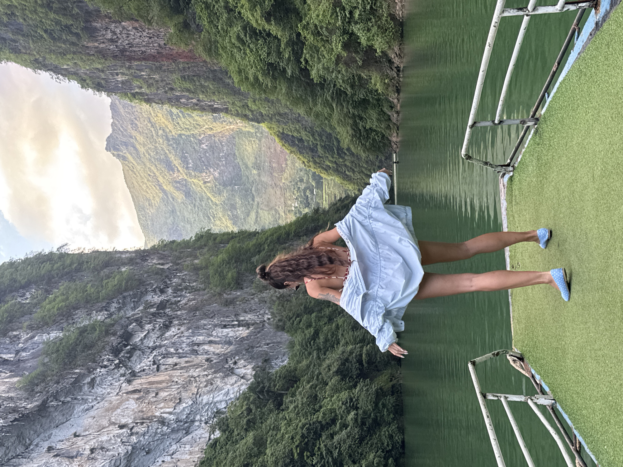 A woman stands on a boat in the city of Đồng Văn Commune, Vietnam. She wears a swimsuit under a light-blue jacket and blue shoes with white polka dots. The boat has a green astroturf floor and white metal railings. In the background, mountains rise on either side of a river. The mountains are covered in green vegetation. The sky is cloudy.