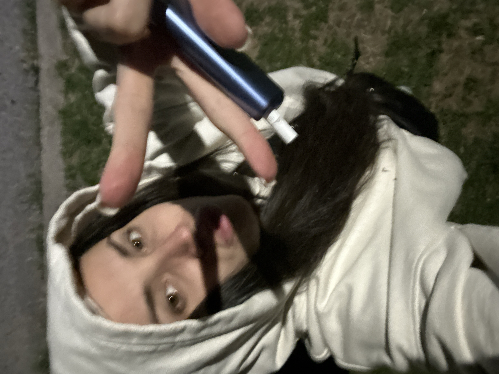 A person is captured in a close-up, low-angle selfie at night. They are wearing a white hooded sweatshirt, with the hood pulled up over their head. Their face is turned towards the camera, with their eyes open and looking at the lens. Their mouth is slightly pursed, and their tongue is sticking out slightly. In their right hand, they are holding a blue cylindrical object, possibly an electronic cigarette device, and have their index and middle fingers extended towards the camera. The background shows a grassy area with some out-of-focus elements that could be pavement or a wall. The lighting suggests artificial light sources are present, creating shadows on the person's face.