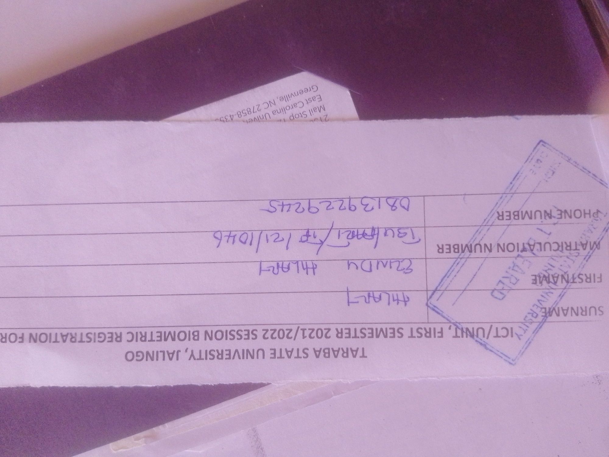 The image is a close-up of a document with handwritten entries and a stamp. The document appears to be a form related to academic registration.
There are several handwritten entries on the form. One entry, written in purple ink, reads "DB139229245". Another entry, in blue ink, seems to be a date, "21/10/121/10/46". Below this, there are names written in blue ink: "EZINDU HILAR" and "AHLAIT".
A blue stamp is visible on the right side of the form. The stamp contains text that is partially obscured. Visible parts of the stamp include "SURNAME", "FIRSTNAME", "MATRICULATION NUMBER", and "PHONE NUMBER". The text "EAST" and "UNIVERSITY" are also visible within the stamp.
The bottom of the document has printed text that reads "ICT/UNIT, FIRST SEMESTER 2021/2022 SESSION BIOMETRIC REGISTRATION FOR TARABA STATE UNIVERSITY, JALINGO".
There is also a piece of paper with printed text that has slid partly under the main document. This text reads "Mail Stop 210 East Carolina University Greenville, NC 27858-435".
The image is taken from a top-down perspective, focusing on the details of the document. The background is a dark, textured surface, possibly a table or a folder, with some purple shading. The lighting is uneven, casting shadows on the document.
The location is identified as the city of Magajin Gari, Nigeria, based on the printed text mentioning "TARABA STATE UNIVERSITY, JALINGO". Taraba State is located in Nigeria, and Jalingo is its capital city, which contains the Taraba State University. Magajin Gari is a district within Jalingo.