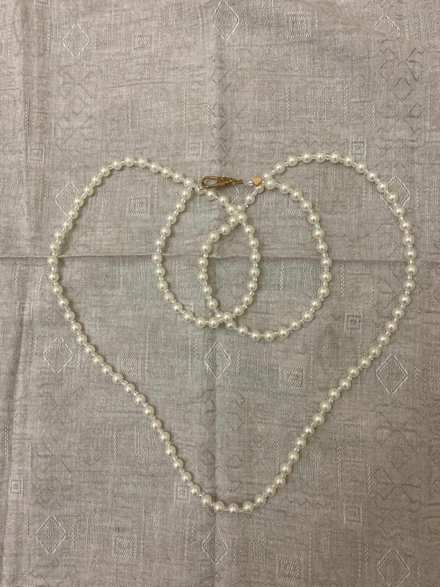 A close-up, overhead shot showcases a pearl necklace with a gold clasp laid out in a heart shape on a light grey, textured fabric. The fabric has a subtle, repeating pattern of geometric shapes, possibly diamonds and squares. The pearls are small and round, with a lustrous sheen, interspersed with smaller, clear beads that catch the light. The gold clasp is visible at the top of the heart shape, connecting the two ends of the necklace. The lighting is soft and even, highlighting the textures of both the pearls and the fabric. The scene is static and appears to be a product shot or a flat lay. The image does not contain any identifiable elements related to a specific location in Islamabad, Pakistan.