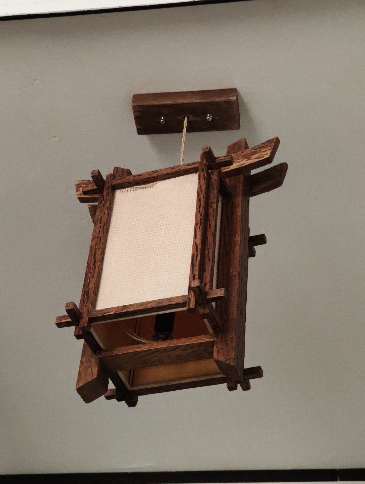 A wooden hanging lamp with a square fabric shade is suspended from a plain, light-colored ceiling. The lamp has a rustic design, with rough-hewn wooden pieces forming its structure. A beige, textured fabric panel is visible within the wooden frame, and an orange-colored material lines the bottom of the lamp. The lamp is attached to the ceiling by a small wooden block and a rope or cord. The background is a neutral ceiling, with a dark horizontal line visible at the very bottom of the frame. The image appears to be an indoor shot taken from a low angle, looking upwards. The location is Komsomolsk-on-Amur, Russia.