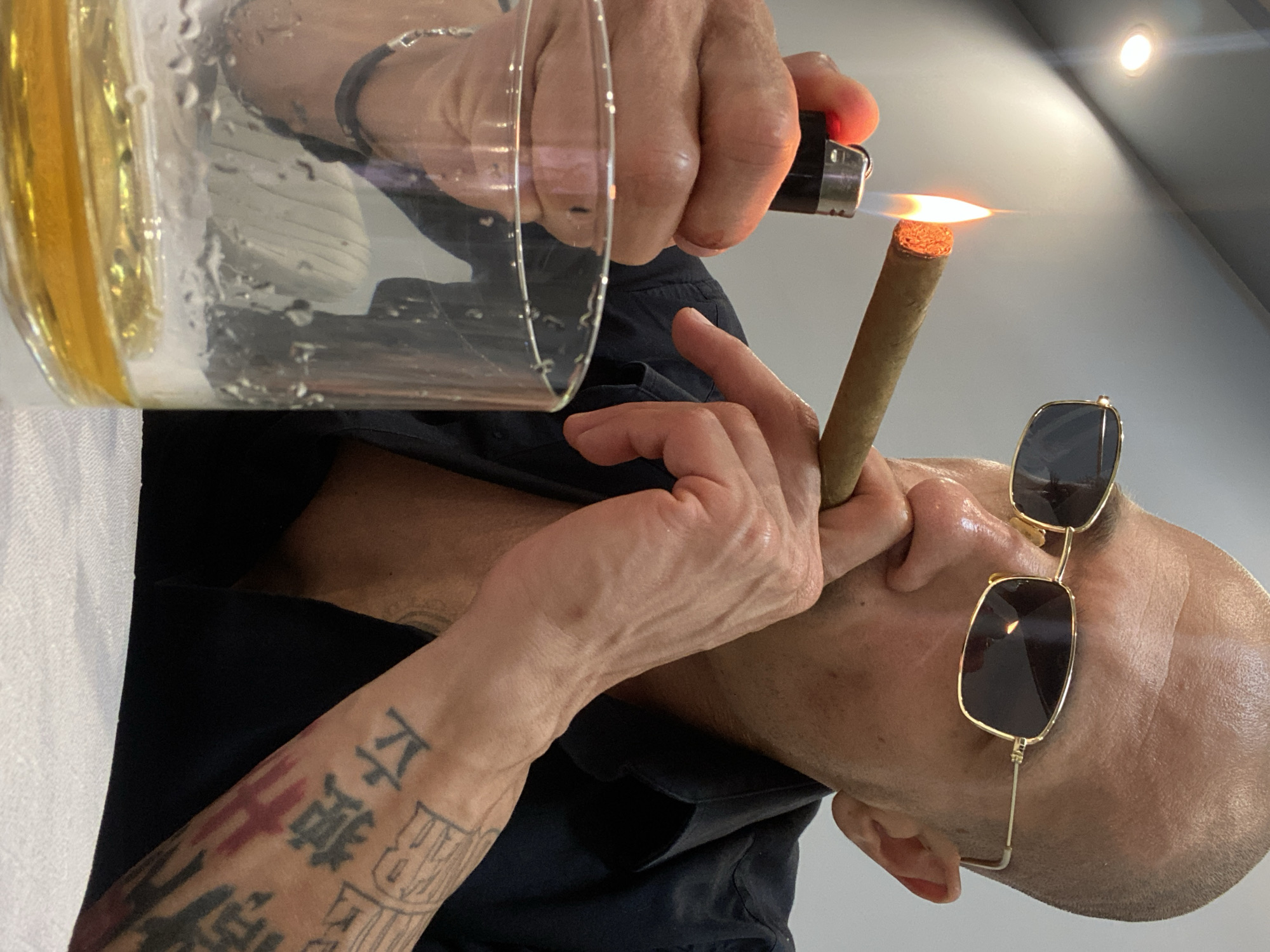 In Pleven, Bulgaria, a man with a shaved head and sunglasses lights a cigar with a lighter. He is wearing a black shirt and has tattoos on his arm. A glass of amber liquid sits on the table in front of him. The scene appears to be indoors, possibly a restaurant or bar, with a muted background. The lighting is warm, highlighting the flame of the lighter and the details of the man's face and hands.