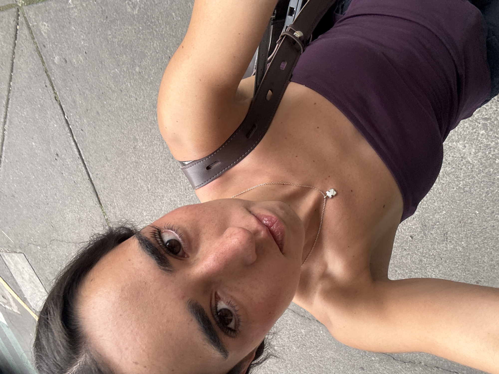 The image is a close-up, high-angle selfie of a person, likely an adult female, taken outdoors in Mexico City, Mexico. The subject is positioned facing upwards towards the camera, which is held above and slightly in front of them, resulting in a somewhat distorted perspective with the head at the bottom of the frame and the body extending upwards to the right.
The person has dark hair, dark, well-defined eyebrows, and prominent eyelashes framing their brown eyes, which gaze directly into the lens. Their lips are full and slightly parted, showing a natural, light color. They wear a dark purple tank top or camisole. A delicate silver-colored chain necklace with a small, white, clover-shaped pendant is visible around their neck. A brown leather shoulder bag strap, featuring visible stitching and buckle details, crosses over their right shoulder and across their chest.
The background primarily consists of grey concrete pavement, suggesting an urban outdoor setting such as a sidewalk or street. The lighting appears bright and even, indicative of daytime, though specific weather like sunny or overcast cannot be determined. The person's expression is neutral to slightly contemplative. No visible text is present in the image.