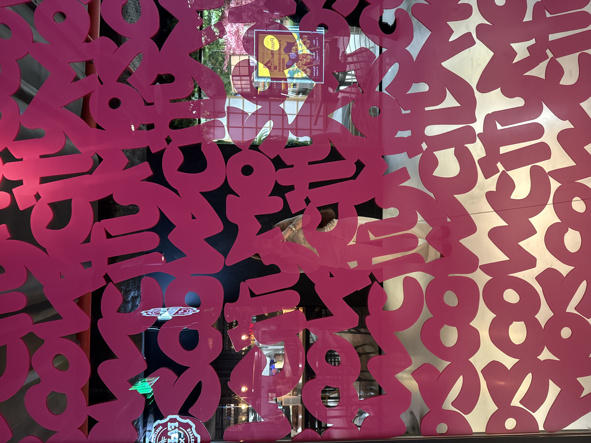 The media file features a large, decorative screen made of opaque, vibrant magenta material, possibly acrylic or vinyl, with numerous abstract, block-like shapes cut out. These shapes appear to be highly stylized letters or numbers, with rounded edges.
Through the cut-out areas and the transparent parts of the glass surface behind the screen, reflections of an interior space are visible. Faint reflections of people can be discerned, along with elements of what appears to be a shop or building interior, including lights and indistinct architectural features.
A notable detail is a square sign reflected in the upper-central portion of the image. This sign displays text predominantly in Cyrillic script. Partially legible words include "Болгарский" (Bulgarian), "ВЕНЫ" (veins, or possibly part of a longer word), and "СЛАДКИЙ" (sweet). Other smaller words like "ОБЕД" (lunch) and numerical figures, likely prices, are also faintly visible on this sign.
In the lower-right corner of the image, a small sticker is affixed to the glass, displaying a logo that includes the English words "BABEL" and "BARBER."
The scene is static, depicting a decorative display rather than an ongoing activity. The bright lighting suggests it could be daytime or a well-lit indoor environment. The presence of Cyrillic text points to a location where this script is in use, such as Eastern Europe or parts of Asia, while the "BABEL BARBER" sticker suggests a commercial establishment like a barbershop nearby or within the reflected space. The dominant colors are the striking magenta of the screen and the muted tones of the reflections.