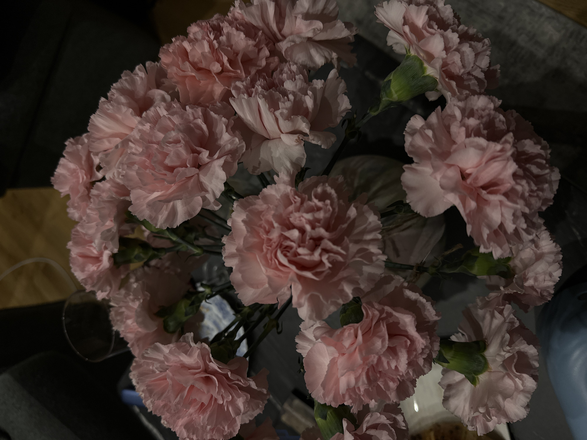 A top-down view of a bouquet of pink carnations in a vase on a reflective surface. The background includes hints of other furniture. No people are visible. The location is unknown, including whether it is in Yerevan, Armenia.
