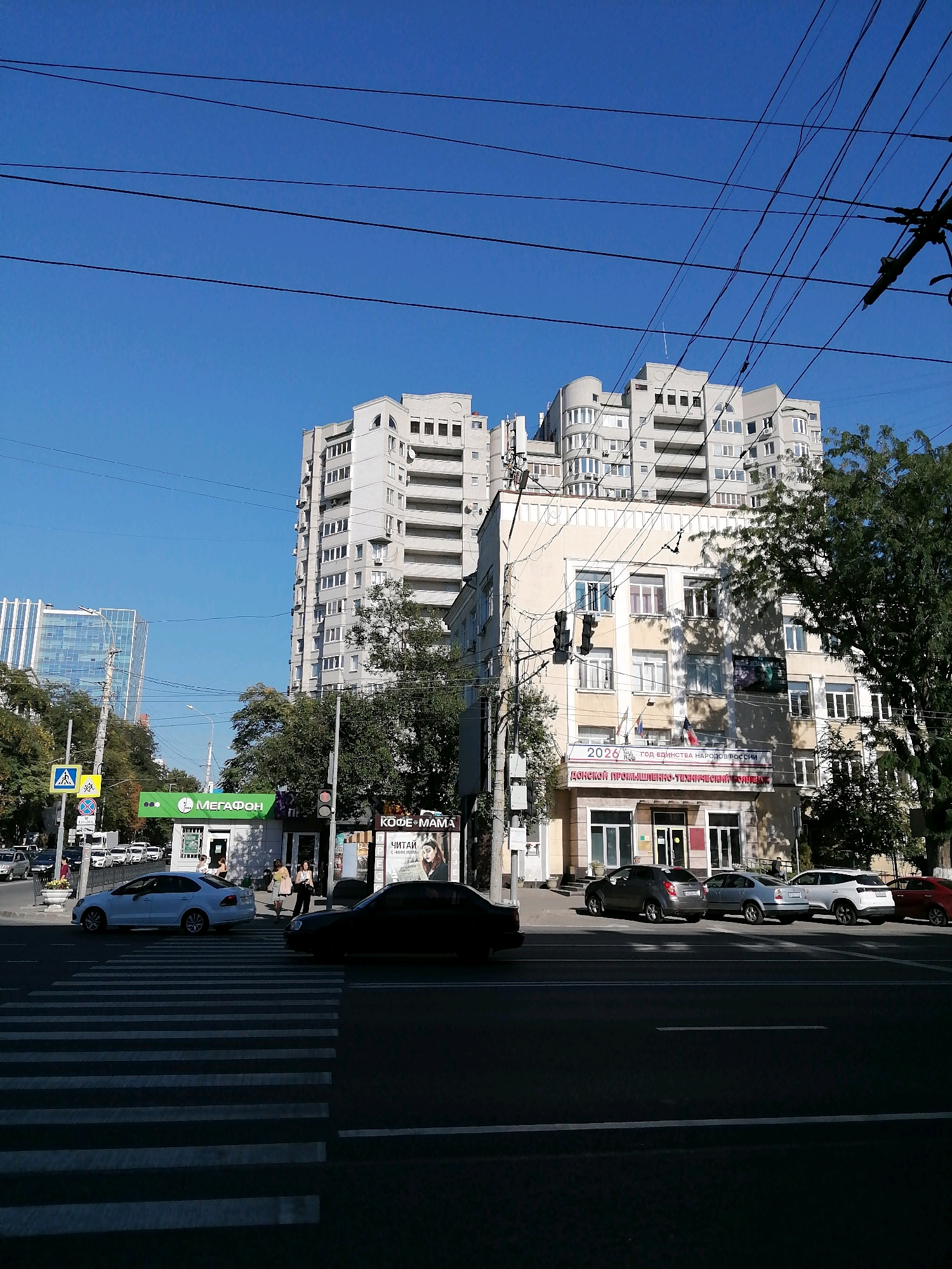 The image depicts a street scene in Rostov-on-Don, Russia, on a clear day. The foreground shows a crosswalk with white stripes on asphalt. Several cars are present: a white sedan stopped at the crosswalk, a dark sedan in the right lane, a gray sedan, a silver sedan, a white SUV, and a red sedan parked on the right side of the street.
Dominating the midground and background are multi-story buildings. To the left, a modern, glass-fronted building stands out, while to the right, a beige building with arched windows features a banner that reads "2026 год Единства народов России" and "ДОНСКОЙ ПРОМЫШЛЕННО-ТЕХНИЧЕСКИЙ КОЛЛЕДЖ" (Don Industrial-Technical College). Above this, another, taller, multi-story building with a curved facade is visible. Another white, block-like building with many windows is also prominent.
Several trees with green foliage are scattered throughout the scene, casting shadows. Electrical wires crisscross the sky. A green kiosk with a "МЕГАФОН" (Megaphone) logo is situated on the sidewalk. People are visible walking on the sidewalk and near the kiosk. Traffic lights and street signs are also present. The sky is a clear, bright blue.