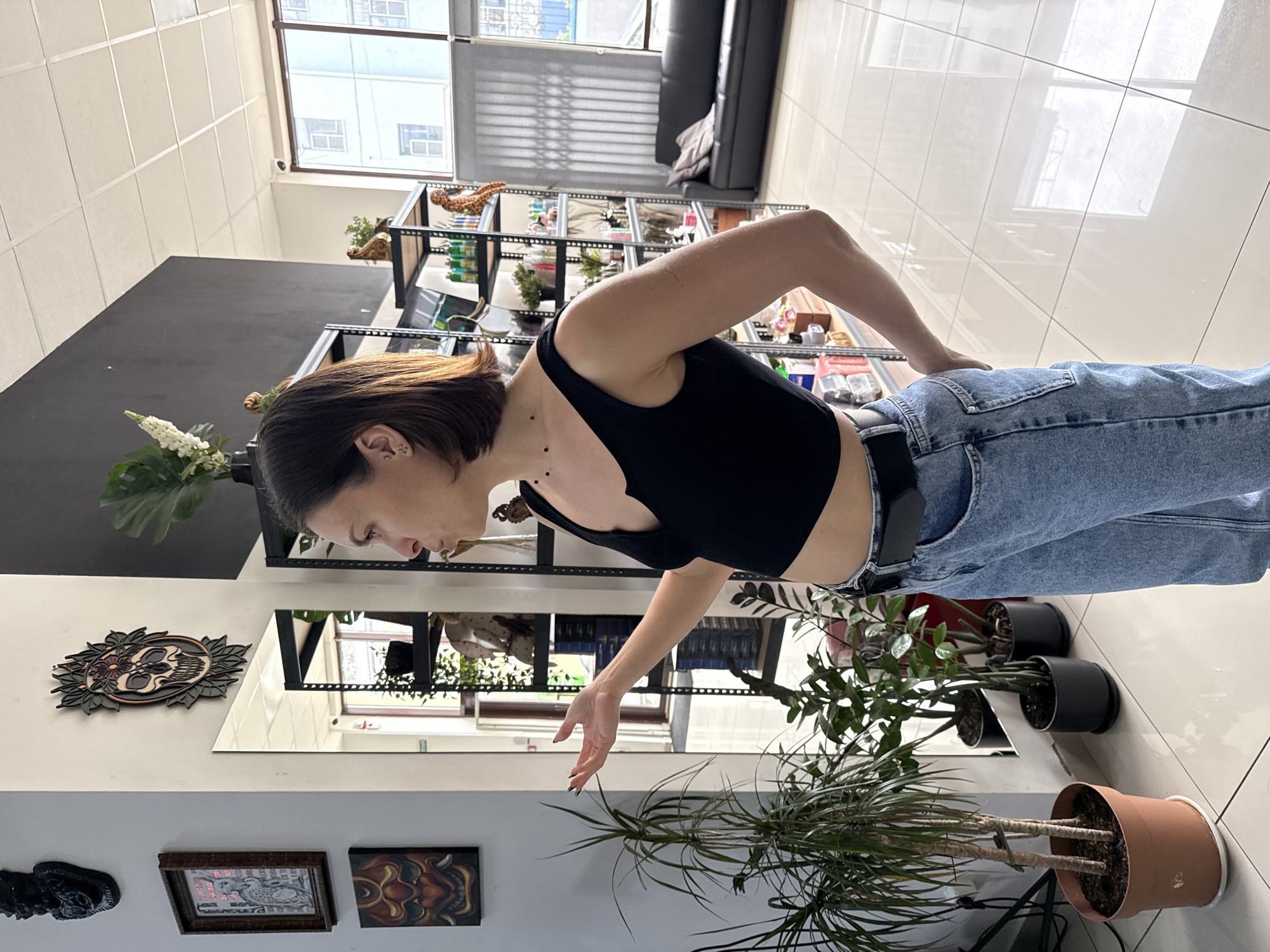 A woman with short brown hair, wearing a black tank top and blue jeans with a black belt, is standing in a room with white tile flooring. She is bending forward with her right arm extended towards a potted plant with long, thin green leaves. Behind her is a black shelving unit with various items on display. The room appears to be a shop or studio, with artwork displayed on a wall visible in a mirror. The location is not explicitly identifiable as Rostov-on-Don, Russia from the image alone.