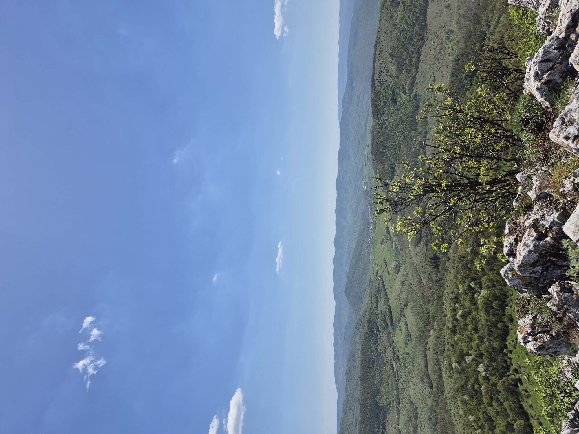 The image shows a landscape view from a high vantage point, likely a cliff or mountaintop, in or near the city of Bilka, Bulgaria. The sky is blue with scattered clouds. Below, a valley or mountainous region is visible, covered in green vegetation, possibly forests and fields. There is a rocky outcrop in the foreground on the right, with a few small trees or bushes growing on it. The overall scene suggests a natural, rural environment with a high elevation perspective.
