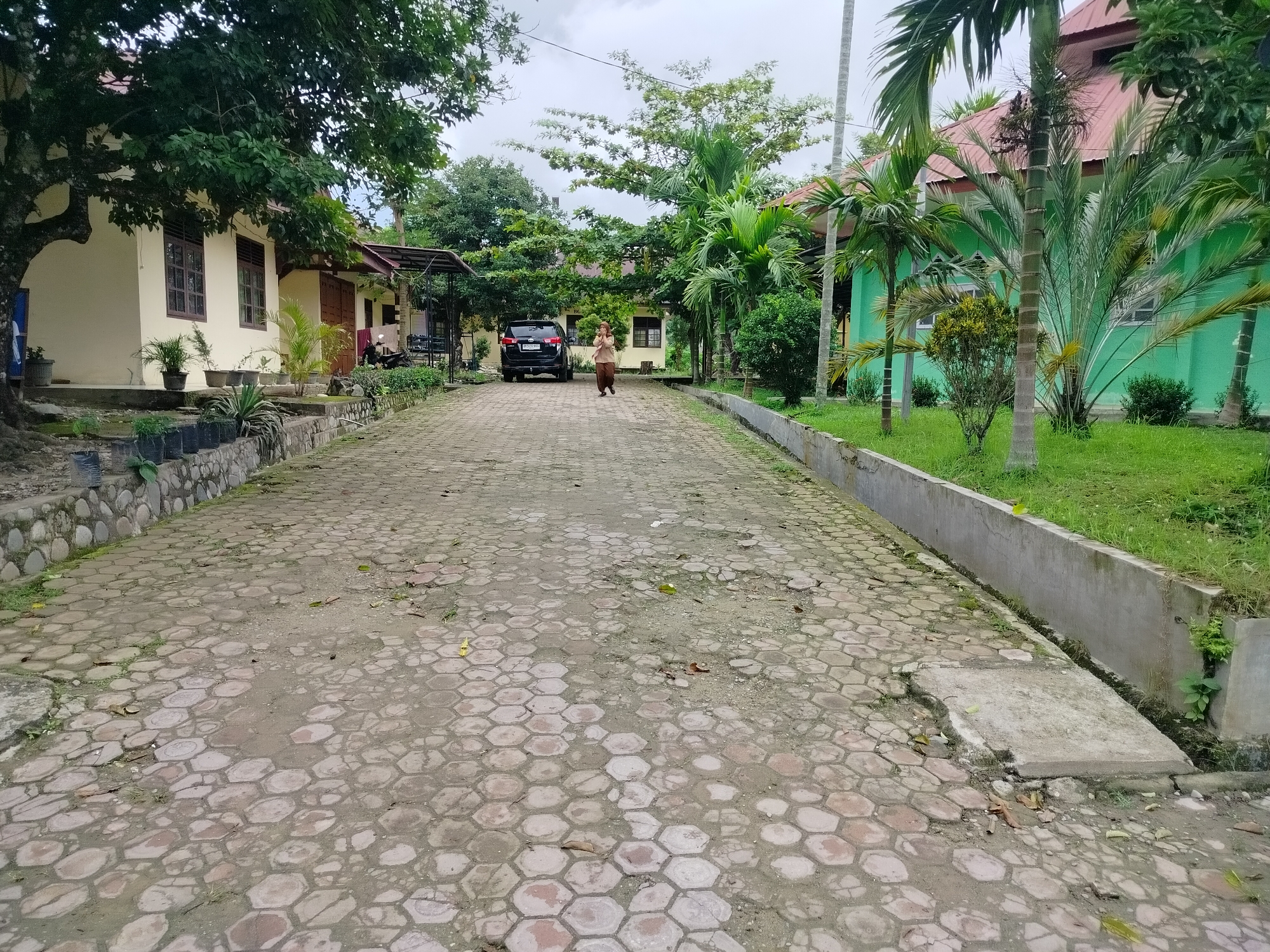 A person walks down a paved driveway in front of light yellow and green buildings. The driveway is made of hexagonal concrete pavers. To the left of the driveway are a light yellow building, a large tree, and a stone wall. To the right are green buildings, palm trees, and a concrete drainage channel. The sky is overcast. The person is wearing a light brown tunic and dark brown pants. A dark SUV is parked on the driveway. The image appears to be taken in Purwodadi, Indonesia, judging by the style of architecture and vegetation.
