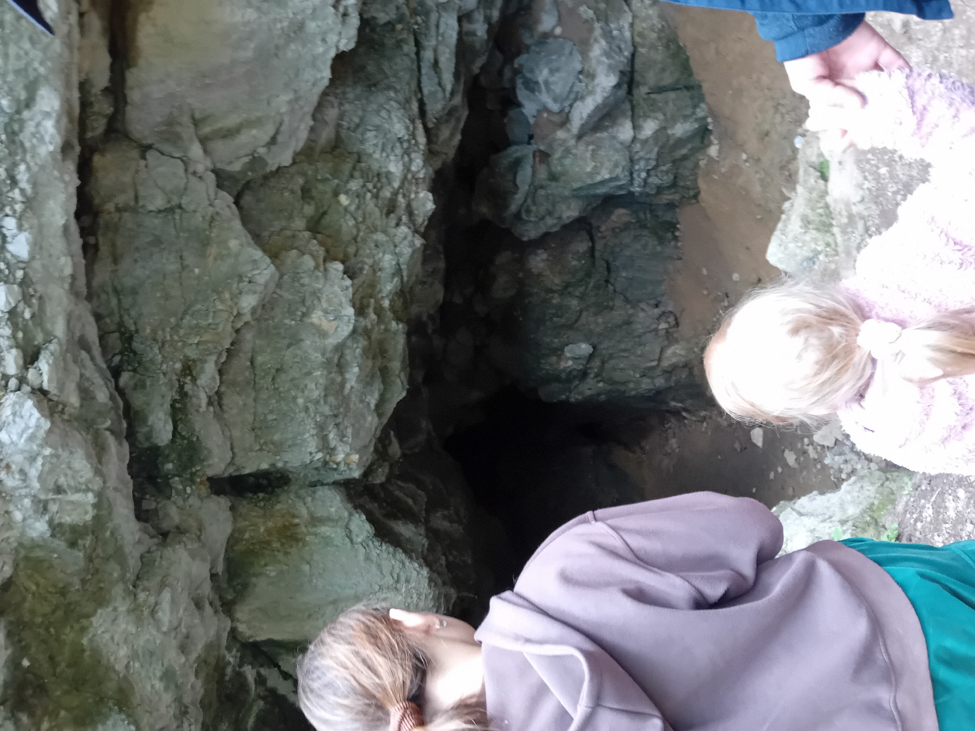 The image shows a cave entrance with a rough, rocky ceiling. In the foreground, two children are visible, facing away from the camera. The child on the left is wearing a purple hoodie and green pants. The child on the right, with blonde hair in pigtails, is wearing a pink fuzzy sweater. A third person's arm and hand, wearing a blue jacket, are visible on the right, holding the hand of the child in the pink sweater. The ground is sandy and rocky. The cave entrance is dark and appears to lead into a cavern. This scene is likely at the Oravice Cave near Zuberec, Slovakia, a natural attraction known for its karst formations.