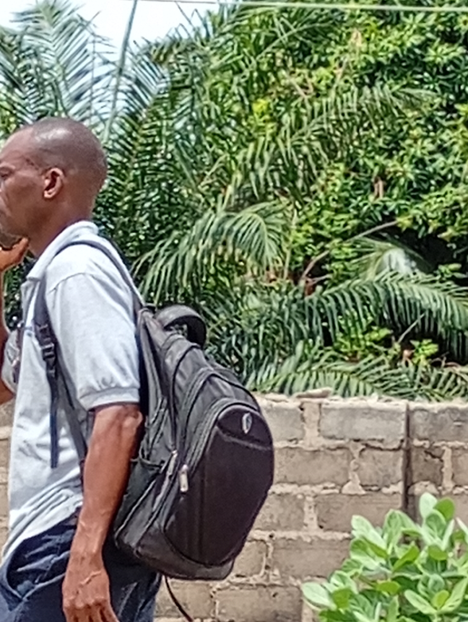 The image shows a man wearing a light-colored, short-sleeved shirt and dark jeans, walking with a black backpack in Lokoja, Nigeria. He is walking past a low brick wall with lush green trees in the background.