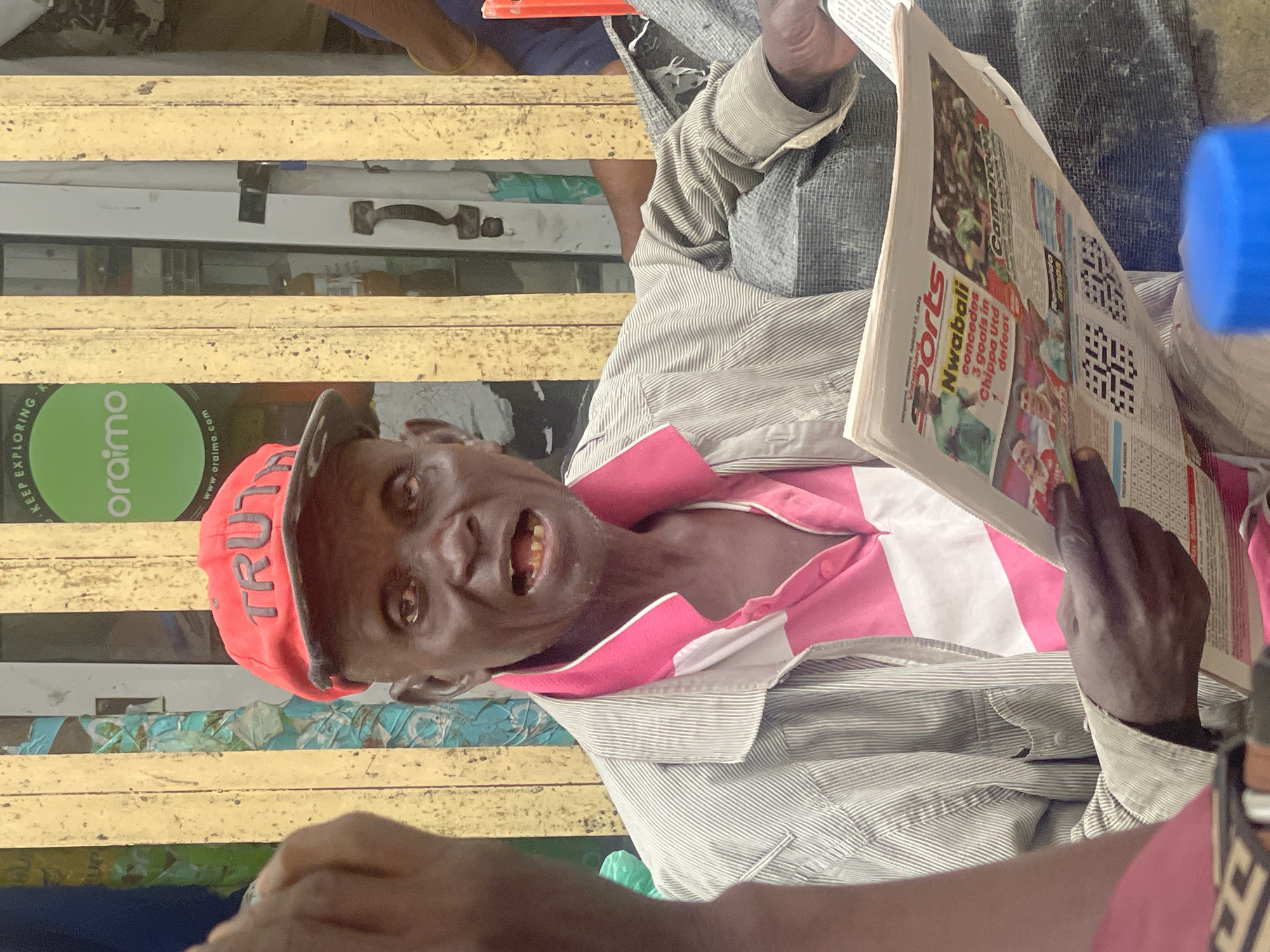 A Black man wearing a red "TRUTH" baseball cap and a pink and white striped shirt is holding a newspaper with the headline "Nwabali concedes 3 goals in Chippa Utd defeat." He is seated outdoors, possibly in a market stall in Lekki, Nigeria, given the style of the wooden posts and the presence of a "Keep Exploring Oraimo" sign in the background, which is a brand often seen in Nigerian markets. The man's mouth is open, and his teeth are visible. The newspaper also features images of soccer players and a crossword puzzle. He is wearing a striped jacket over his shirt. The overall scene depicts a street vendor or resident engaged with daily news.