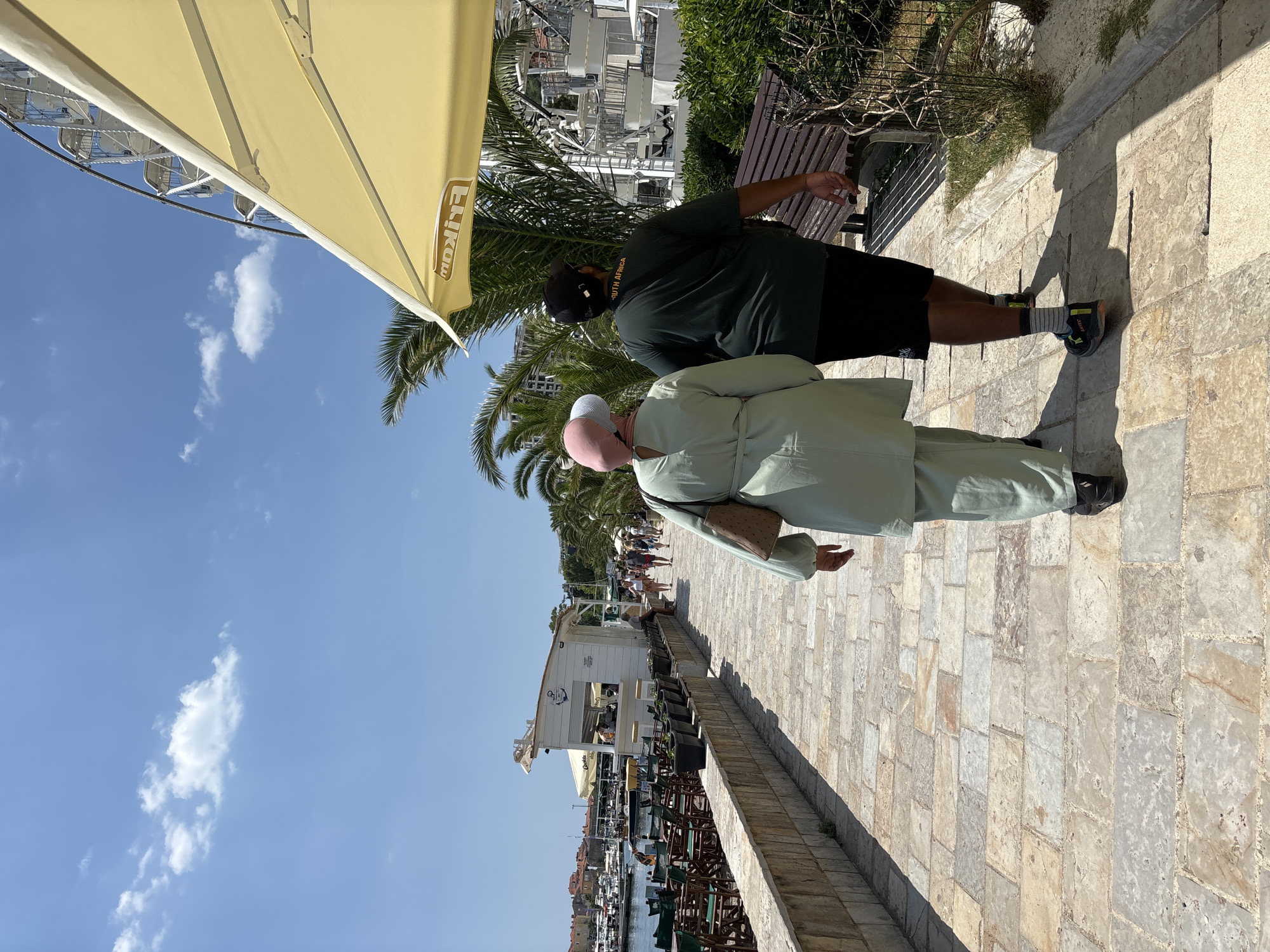 Two individuals, one wearing a light green abaya and pink hijab, and the other a dark green t-shirt and black shorts, walk away from the camera on a stone-paved promenade. They are walking past a row of potted plants and a low stone wall on their left, which separates the walkway from a body of water with boats docked. On the right side of the promenade, there are palm trees, bushes, a park bench, and a Ferris wheel in the background. The scene appears to be in Budva, Montenegro, likely near the marina. The sky is clear blue with a few white clouds.