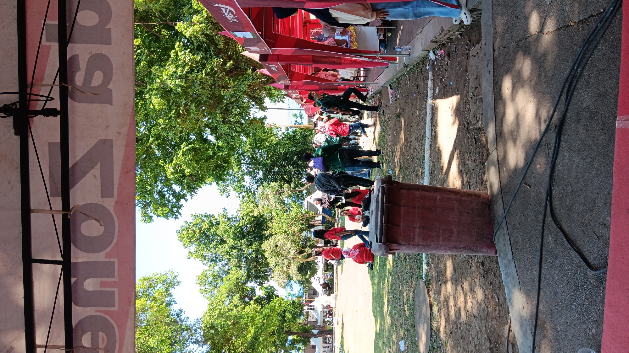 The image depicts an outdoor gathering, possibly a community event or market, in Nganjuk, Indonesia. The scene is characterized by numerous red tents set up in rows, suggesting a commercial or exhibition area. Several people are visible, some interacting near the tents, while others are scattered across a grassy area. A large, reddish-brown trash bin is prominently placed in the foreground on the left. The environment is filled with lush green trees, indicating a park-like setting or a location with ample natural foliage. The ground appears to be a mix of dirt and paved surfaces, with some cables visible on the ground in the foreground. The overall atmosphere seems casual and social.