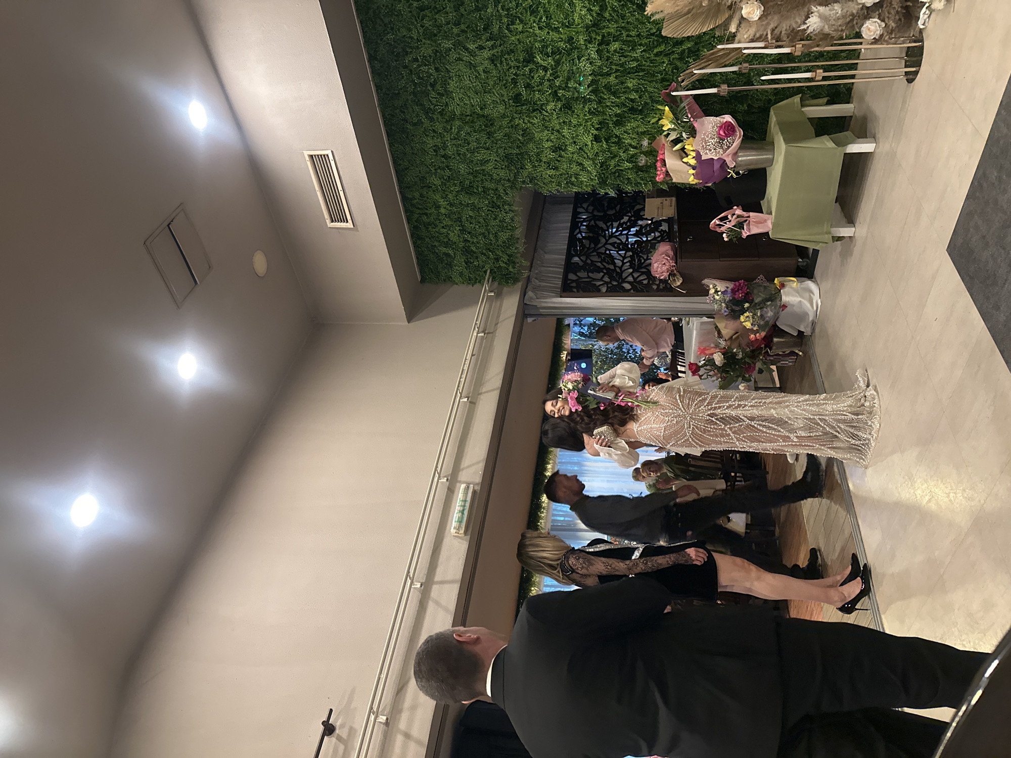 A photo shows a group of people standing in an indoor event space. The setting includes a wall decorated with artificial greenery, suggesting a modern or upscale venue. A woman in a long, ornate dress stands at the center, likely the focus of attention. Other people are positioned around her, dressed in formal wear, indicating the event is formal. There are floral arrangements and decorative elements. The lighting in the room appears to be artificial. The location is unspecified in Sofia, Bulgaria.