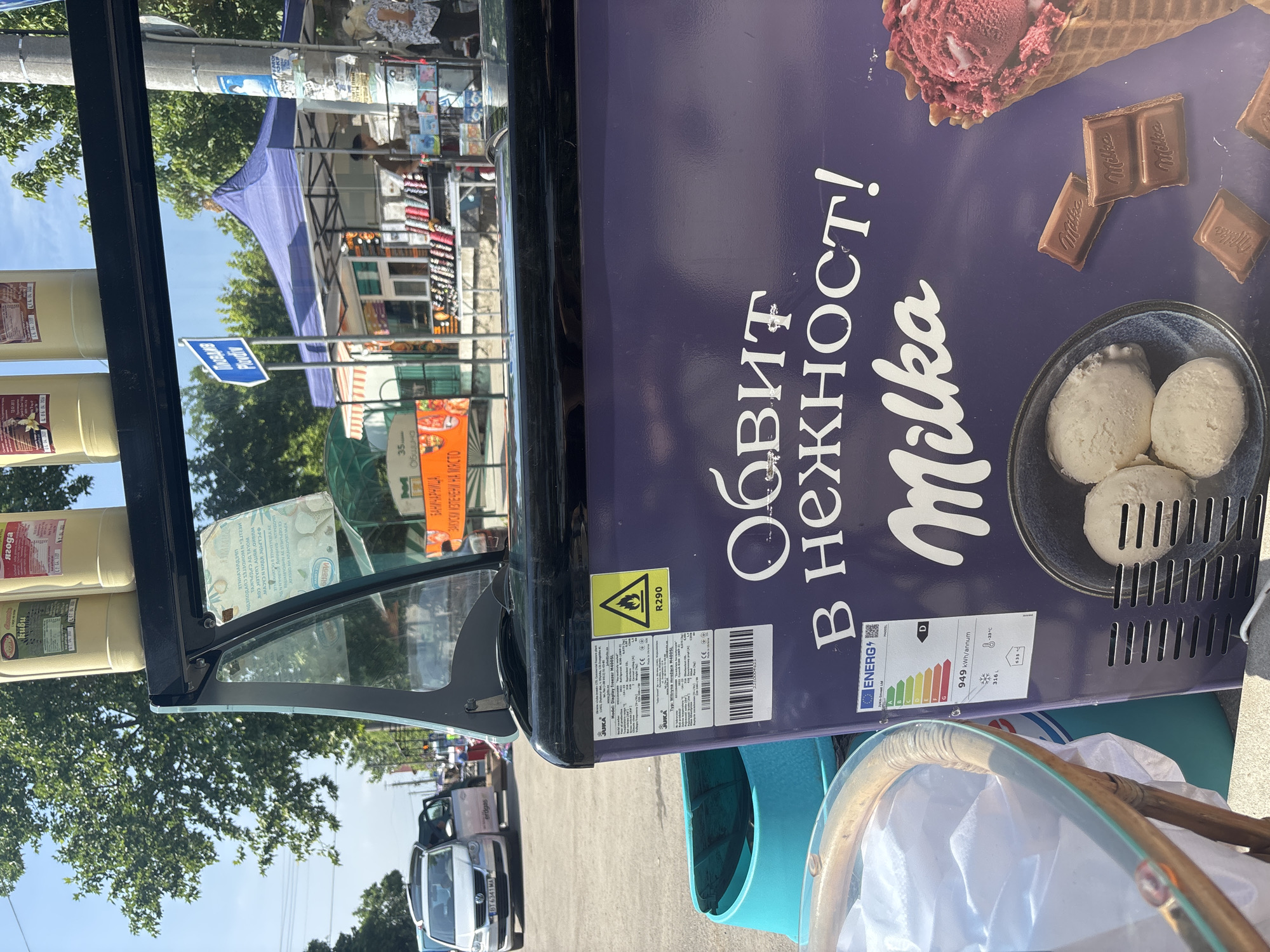This detailed description portrays an outdoor street scene, likely a market or commercial area, under bright daylight conditions. The primary subject in the foreground is a purple, Milka-branded ice cream freezer with a partially open glass lid, angled slightly to the right. The freezer features large white Cyrillic text reading "ОБВИТ В НЕЖНОСТ!" (Wrapped in tenderness!) and "Milka". An energy efficiency label (rated D, consuming 949 kWh/annum) and a technical label specifying "JUKA" vertical freezer M400SL, using R290 refrigerant, are affixed to its side. The freezer's facade also displays images of three scoops of vanilla ice cream in a dark bowl, several pieces of Milka chocolate, and a red berry-flavored ice cream cone. Two cream-colored plastic bottles rest on the black frame above the freezer. To the immediate left of the freezer, a light turquoise plastic bin is visible, alongside a small, round table with a glass top and a wicker-like edge, covered with a white cloth or bag.
In the midground, beyond the freezer, an asphalt street is visible. A silver Volkswagen van with Bulgarian license plate "BT 6341 MA" is parked on the left, with its passenger door open. Further back, a vibrant market setting unfolds. A prominent blue tent-like canopy covers a stall. To its left, a light green awning shades another vendor. A tall utility pole, extensively covered with various papers and posters, stands to the right of the blue tent. A blue rectangular directional sign on a pole, reading "Поща Plovdiv" (Post Office Plovdiv), is visible behind the market stalls. Another sign for a "Баничарница" (Bakery) with text "ЗАКУСКИ ИЗПЕЧЕНИ НА МЯСТО" (Snacks baked on site) is partially visible on the far right. Figures of people can be seen interacting with the market stalls; one individual with a light-colored hat is visible near the utility pole.
The background features numerous dense green trees under a clear blue sky. The overall impression is that of a bustling local market street in Kostievo, Bulgaria, on a sunny day.