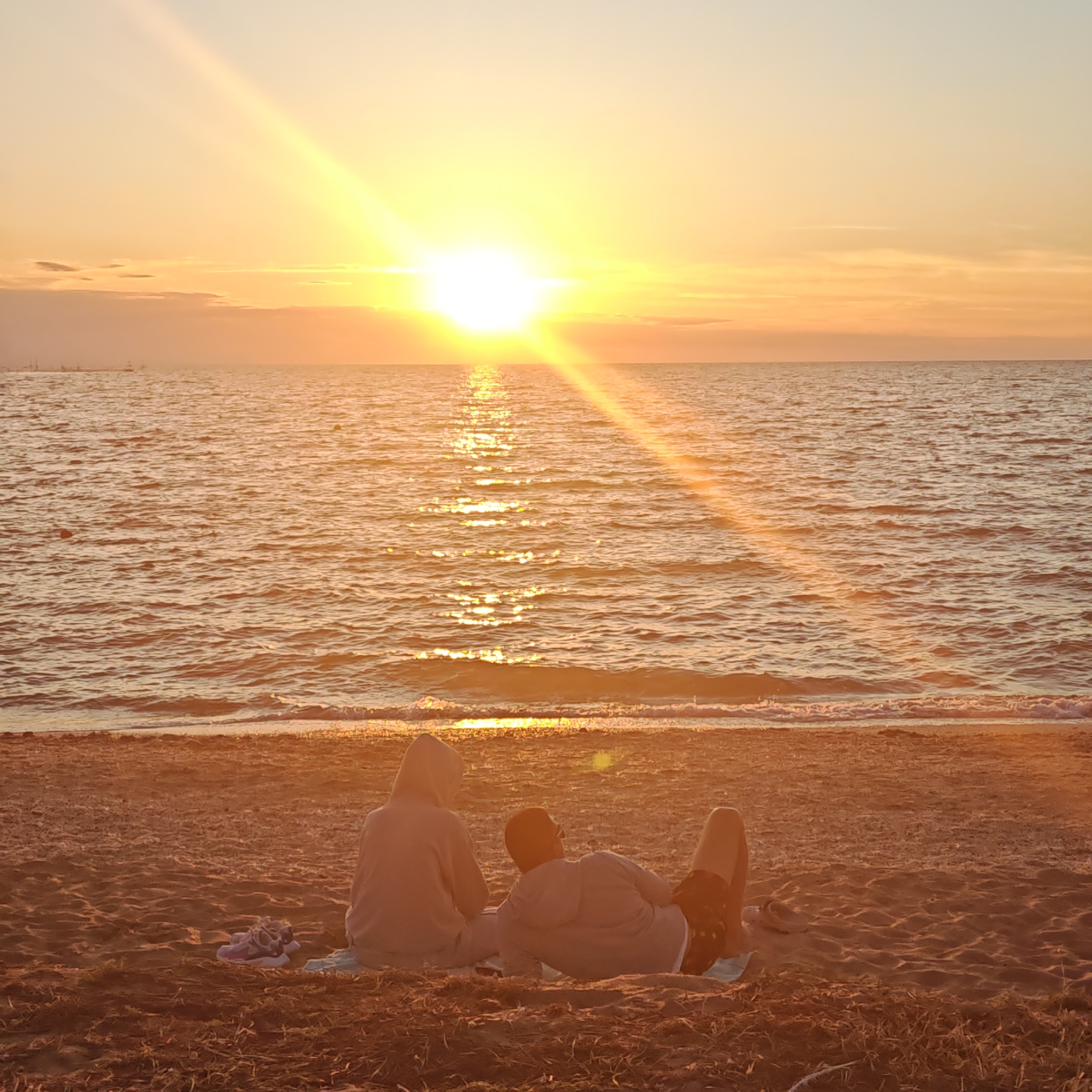 Two people are sitting on a sandy beach facing the ocean at sunset. The person on the left is wearing a light gray hooded sweatshirt and is seated with their legs extended. The person on the right is wearing a light gray hooded sweatshirt and patterned shorts, and is reclining on their side with their legs bent. A small dog is lying between them. A pair of sandals are on the sand to the left of the seated person. The sun is setting over the ocean, casting a warm glow and creating a shimmering reflection on the water. The sky is a gradient of orange and yellow, with some clouds. The beach is sandy with some dried grass and debris.