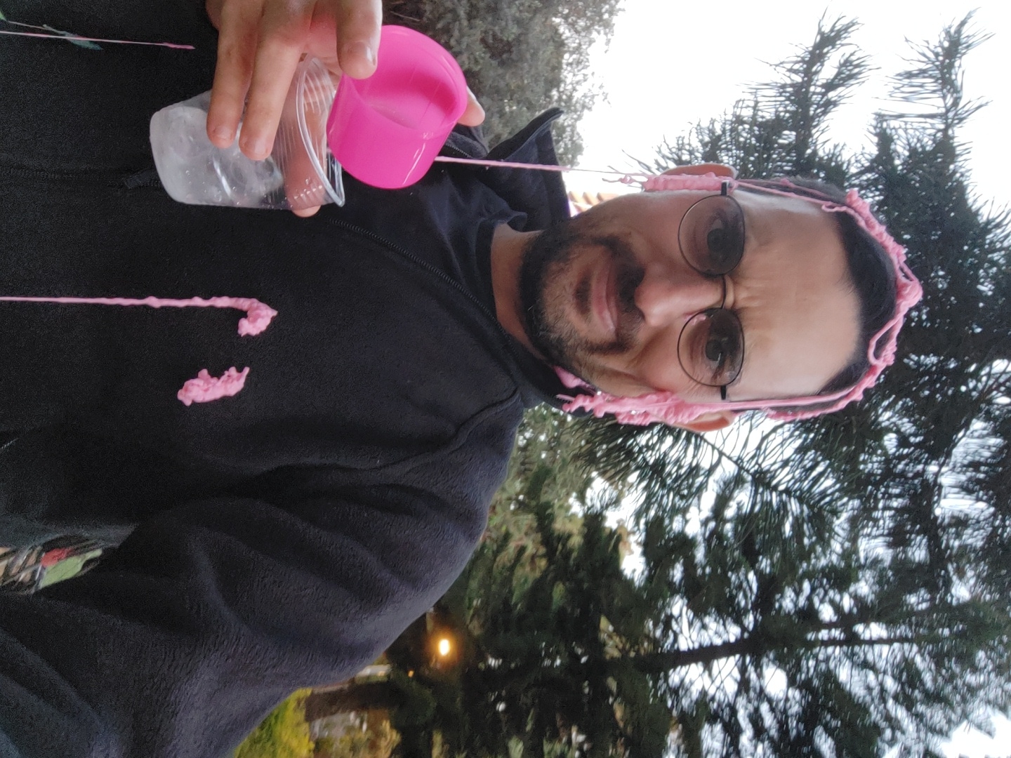 A man with a beard and round glasses is captured in a playful outdoor scene. He is wearing a dark, possibly black, hooded fleece jacket. Pink, viscous, and stringy material, resembling slime or a similar substance, is draped over his head, glasses, and ears, and is also splattered on his jacket. His right hand is visible, holding a clear plastic cup containing ice cubes, while a pink plastic cup is positioned above it, from which the pink substance appears to be pouring or has just been poured.
The man's facial expression suggests a mix of surprise, amusement, and a slight grimace, indicating a reaction to the messy situation. He is looking directly at the camera.
The setting is outdoors, with tall, coniferous trees (possibly pine trees) visible in the background, against an overcast or bright sky. A faint light source, possibly a distant lamp, can be seen in the lower right portion of the background, suggesting it might be late afternoon or dusk. No visible text is present in the image. The location context provided is Tel-Aviv, Israel.
